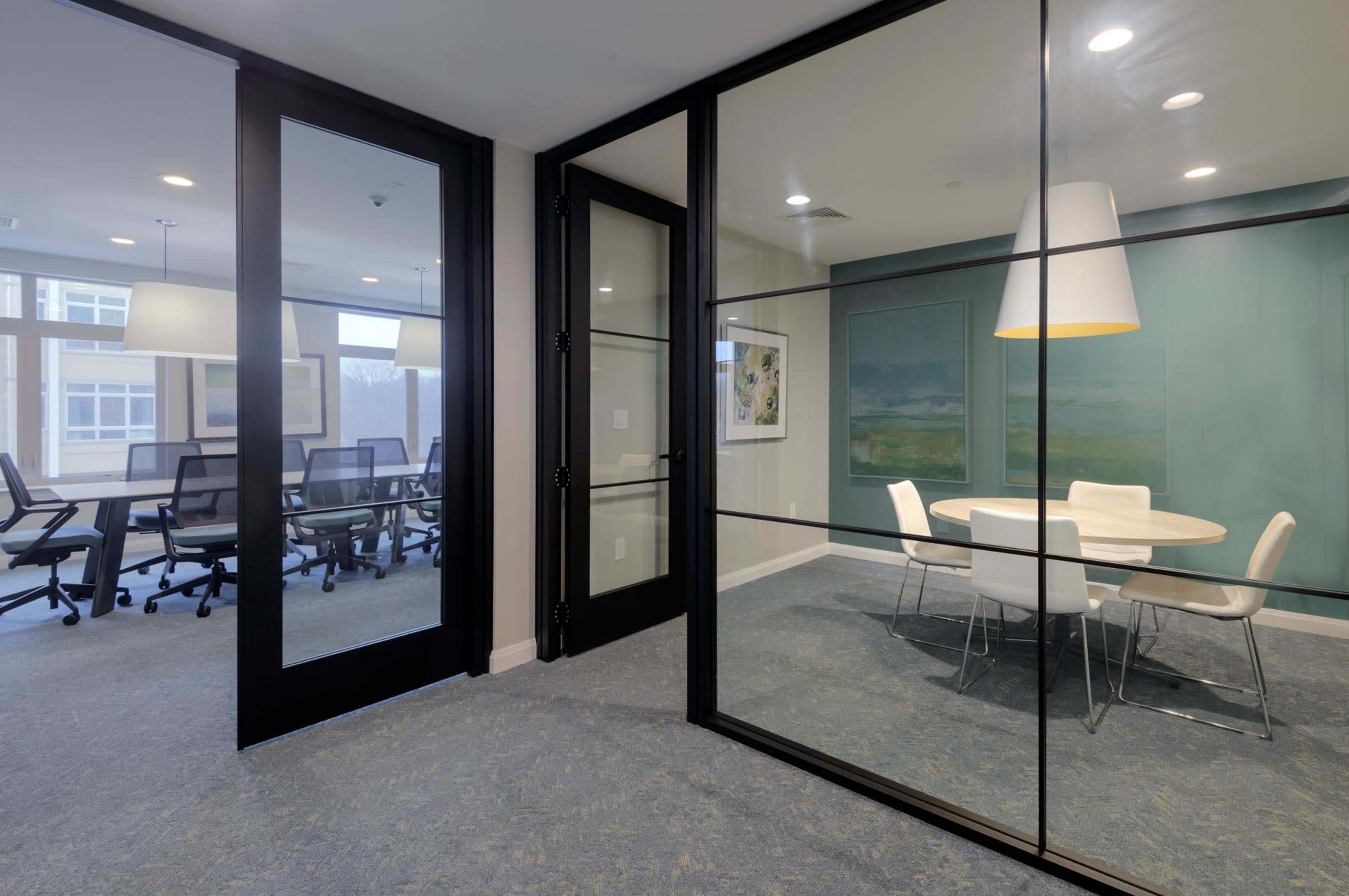 A modern office space with glass walls separating two meeting areas, one featuring a round table and white chairs and the other with conference room seating.