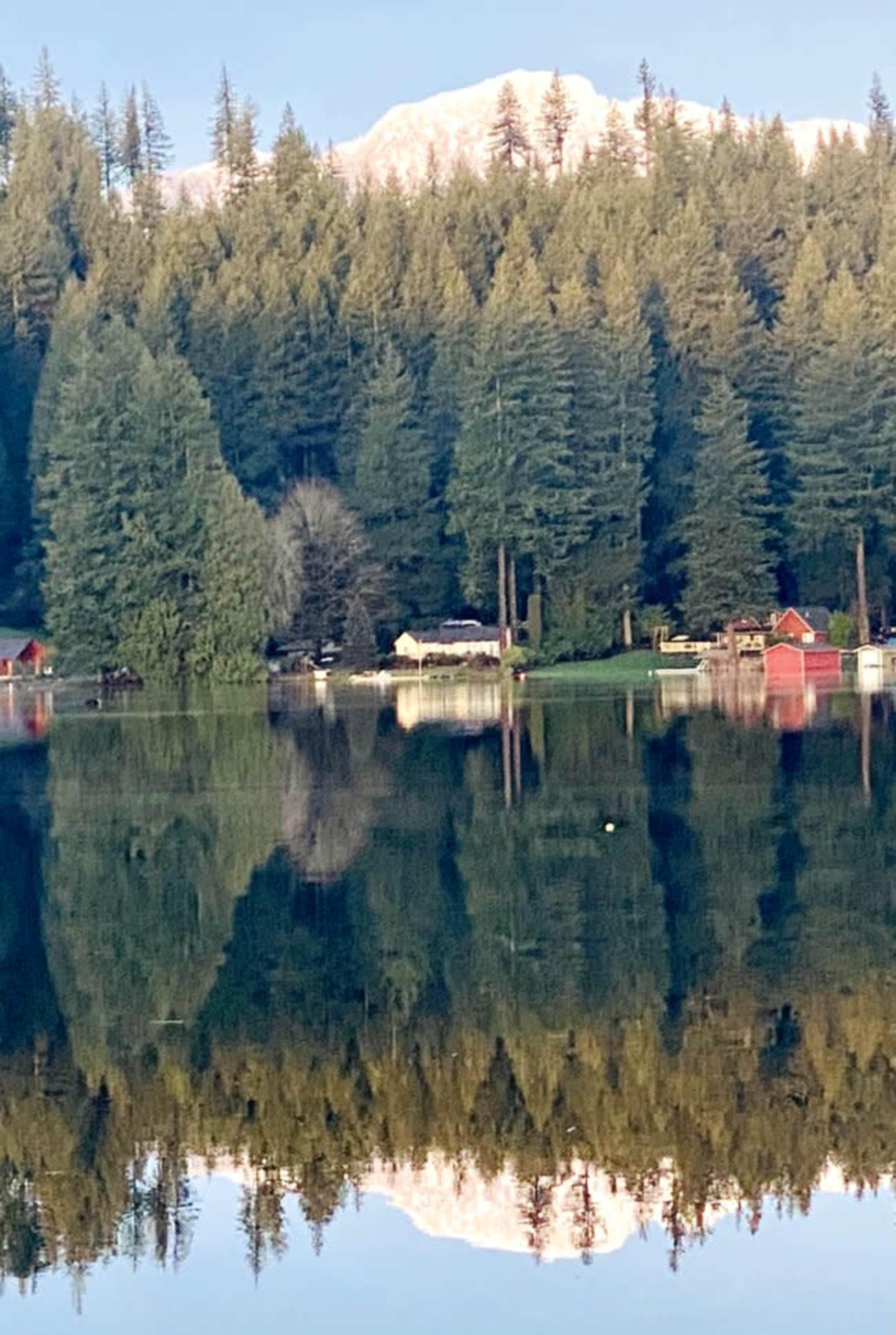 A calm lake reflects a row of evergreen trees and a snow-capped mountain in the background, with a few red cabins visible along the shore.