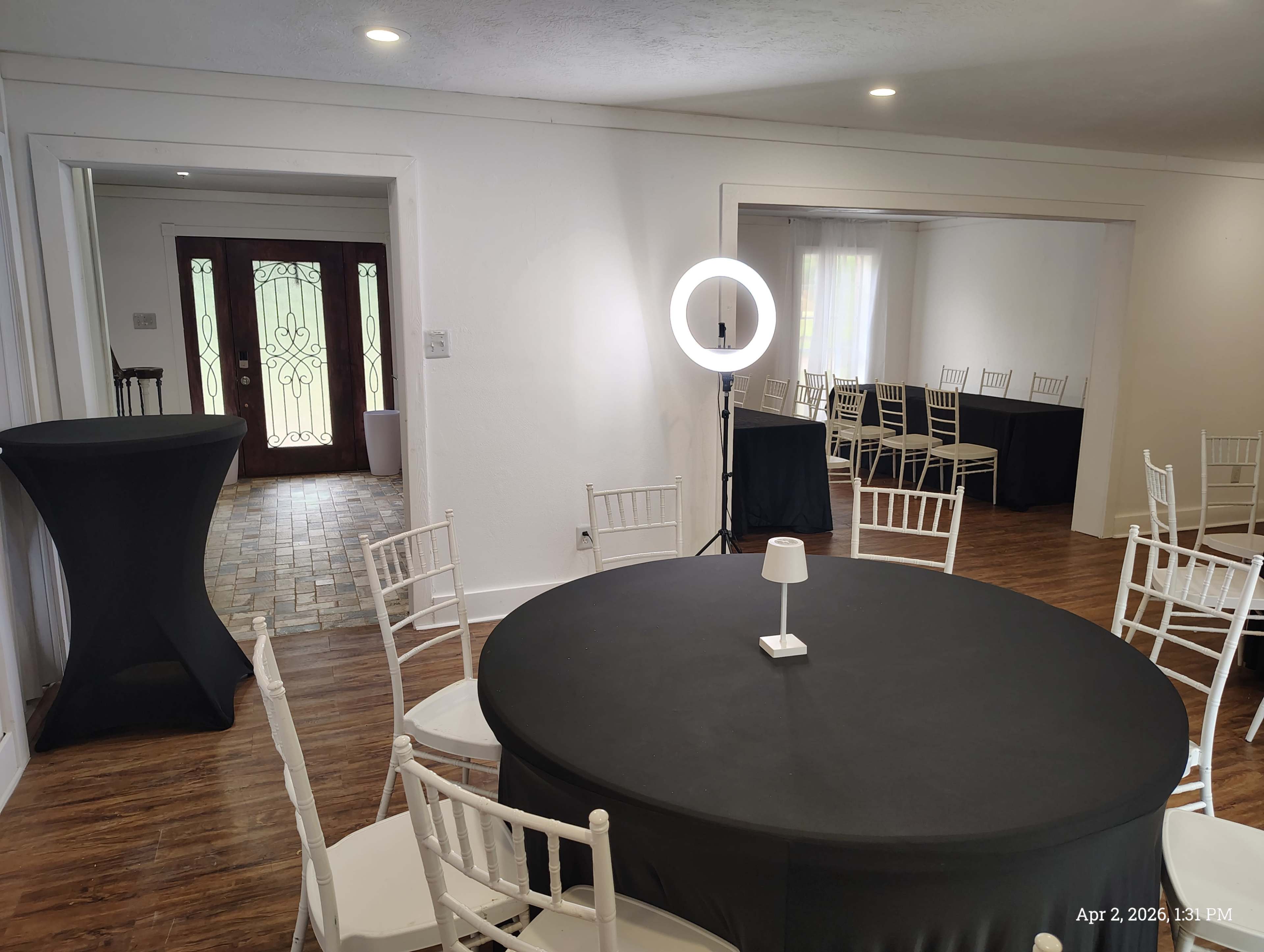 The image shows a spacious room set up for an event, featuring a round table covered with a black tablecloth, surrounded by white chairs, and a backdrop of a doorway with decorative glass.