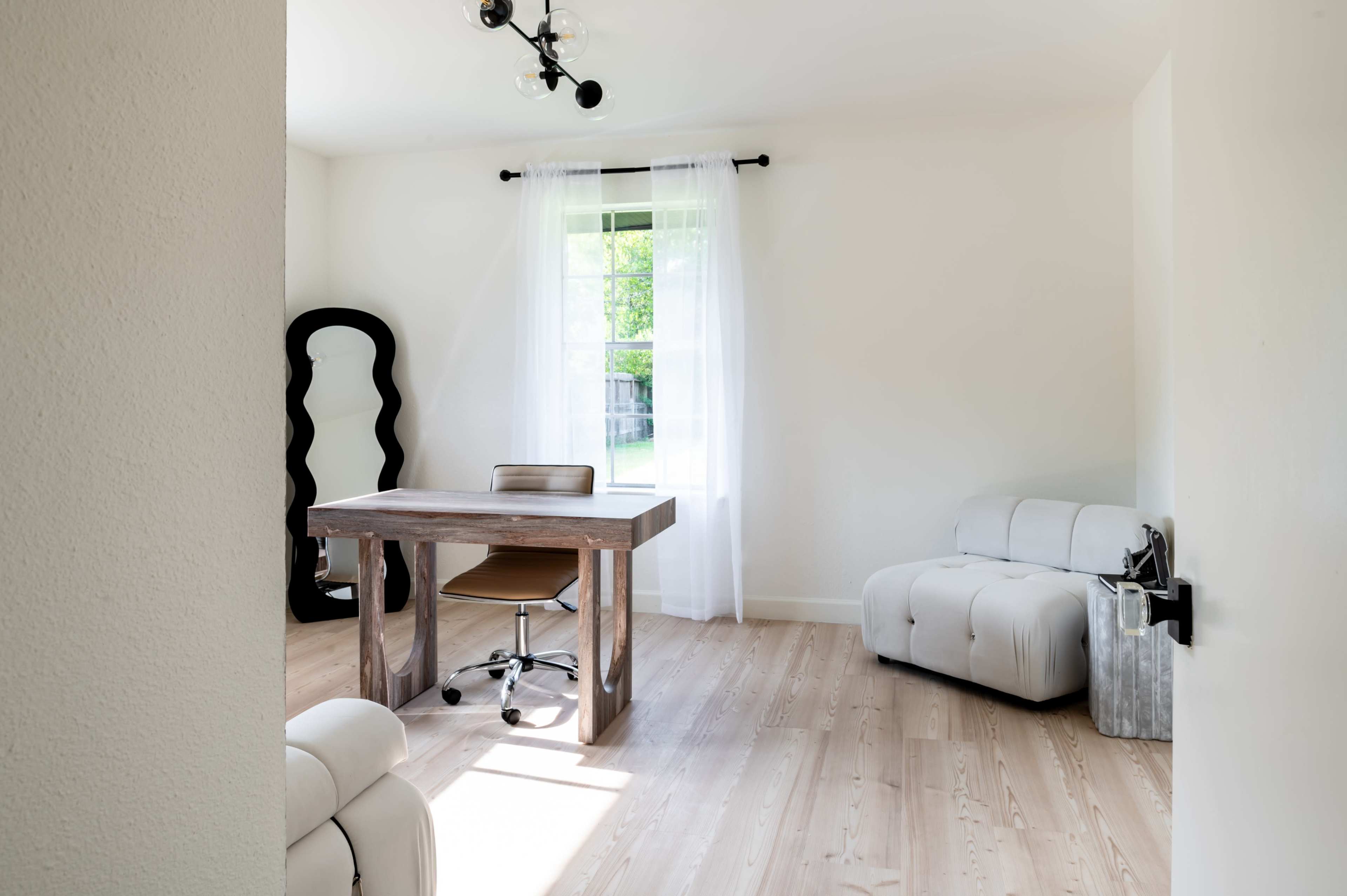 A minimalist home office features a wooden desk, a chair, and a light-colored couch, with natural light illuminating the space through a window.