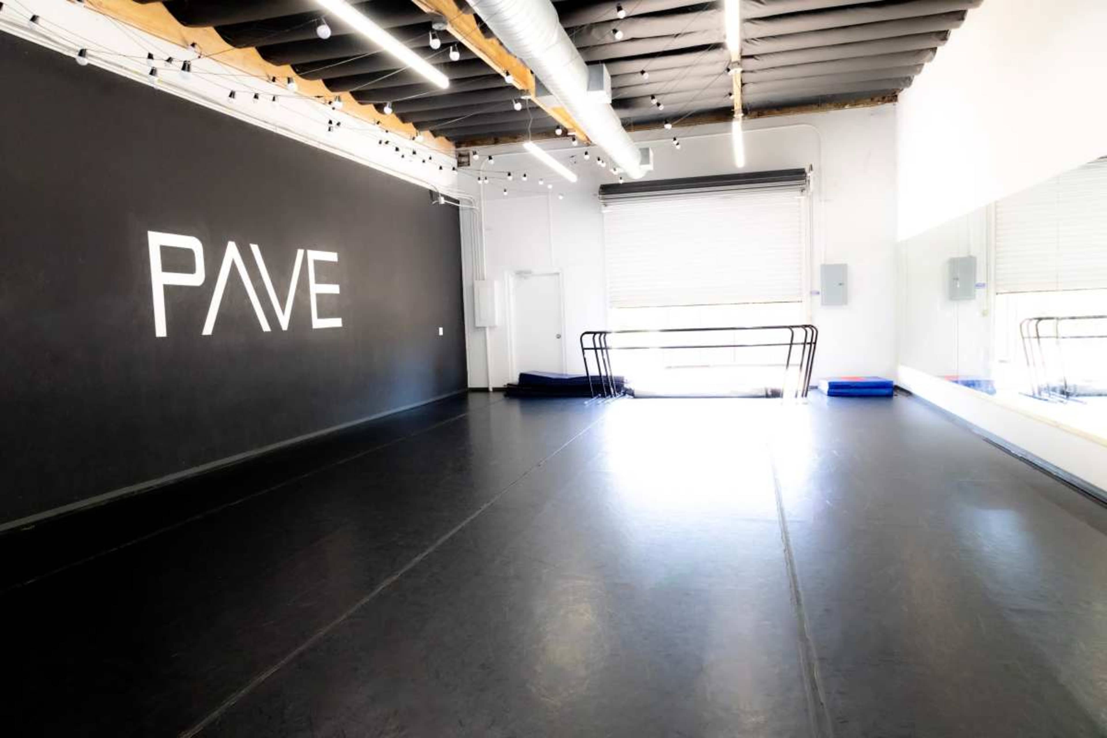 The image shows a spacious dance studio with a mirrored wall, a black floor, and the word "PAVE" displayed prominently on the back wall.