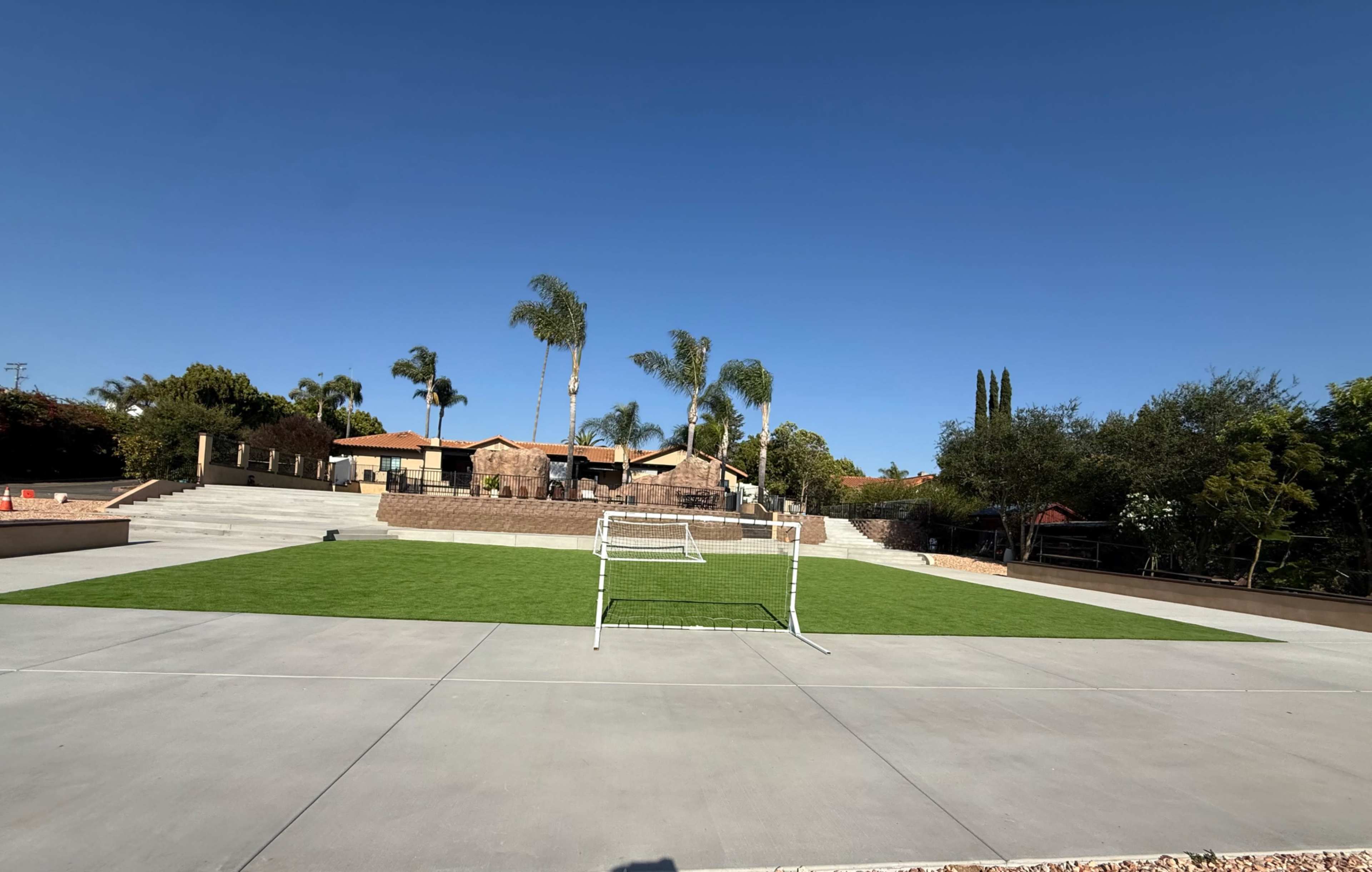 The image shows a well-maintained grass soccer field with a pair of goals in front of a landscaped area featuring palm trees and a residential backdrop.