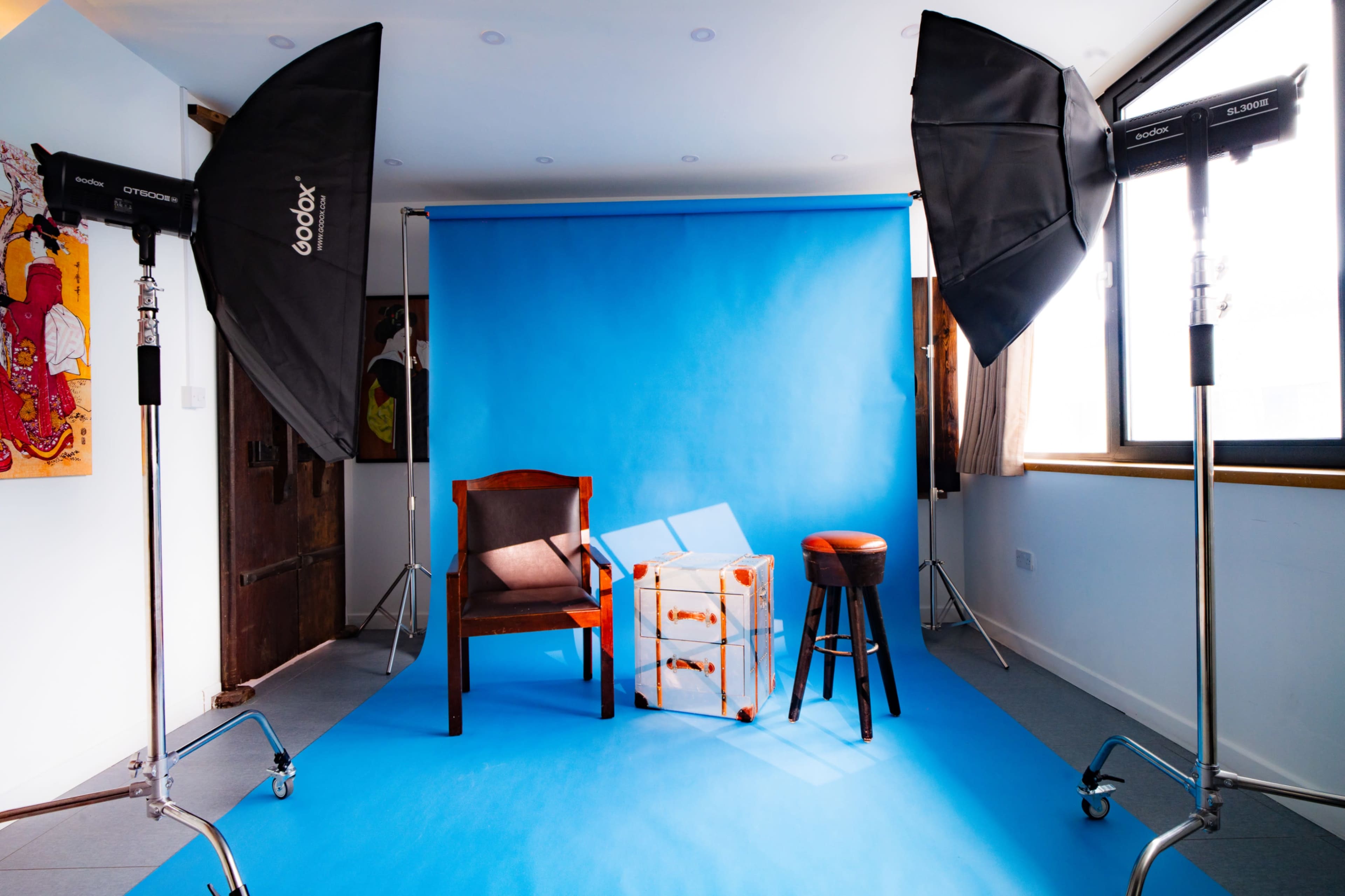 The image shows a photography studio setup featuring a blue backdrop, two large softbox lights, a wooden chair, a stool, and a suitcase on a blue floor.