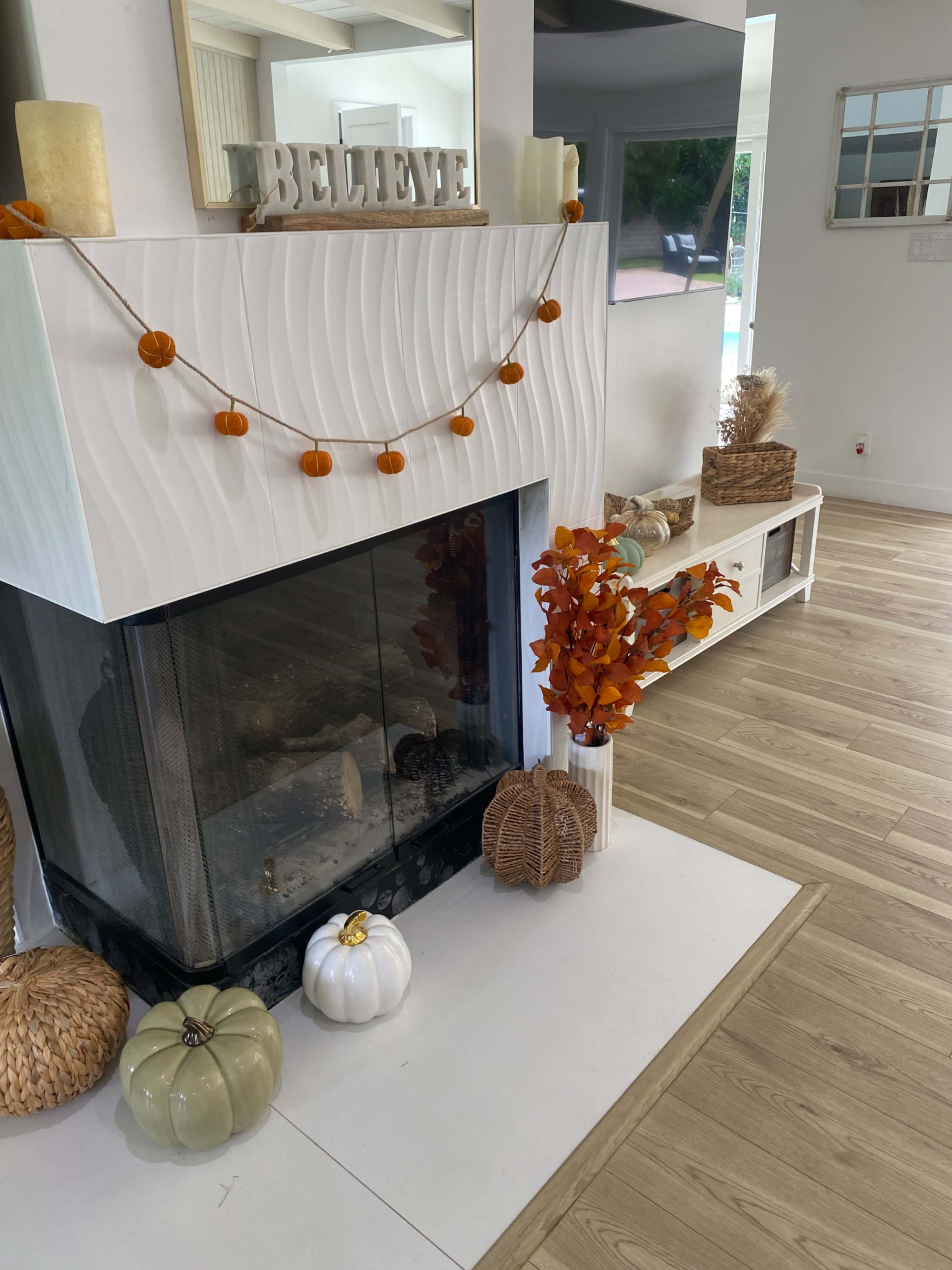 A fireplace with a wavy white facade is adorned with a pumpkin garland, and decorative items include faux pumpkins and a vase of autumn leaves.