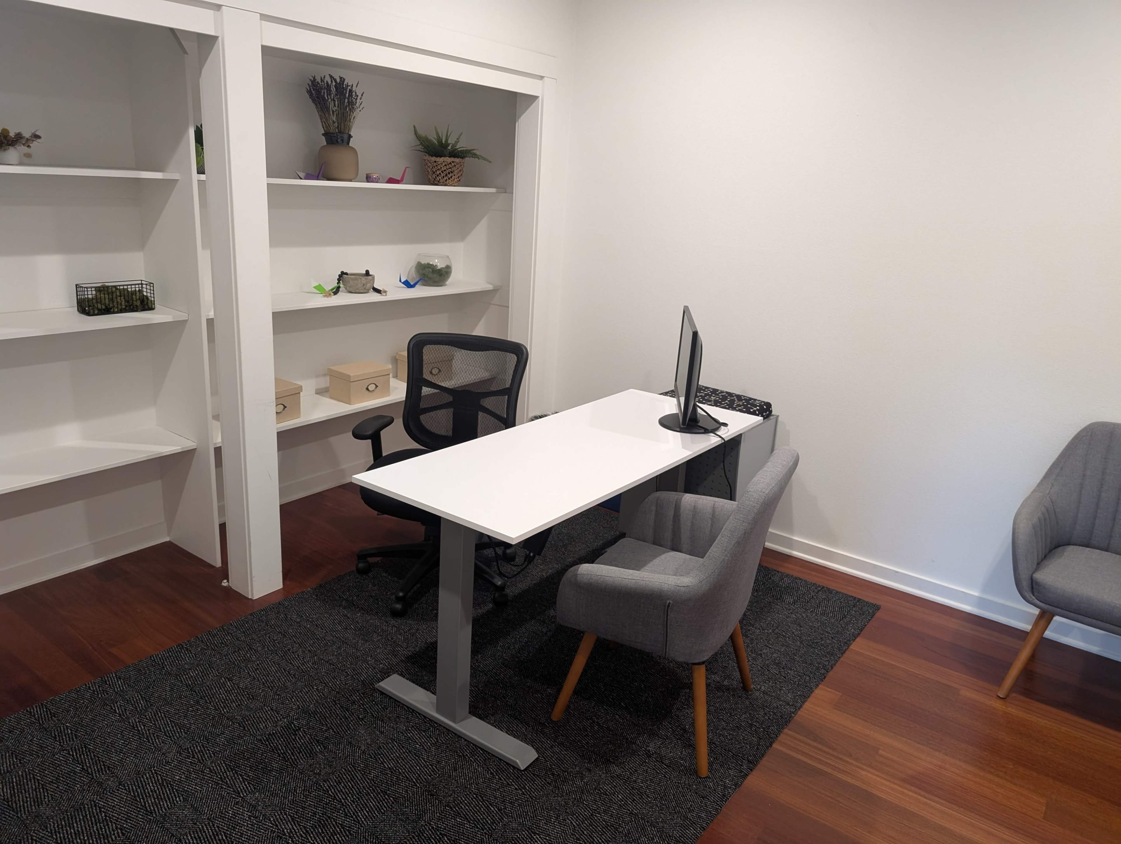 The image shows a minimalistic office space featuring a white desk with a computer monitor, a black office chair, and two gray chairs on a dark rug.