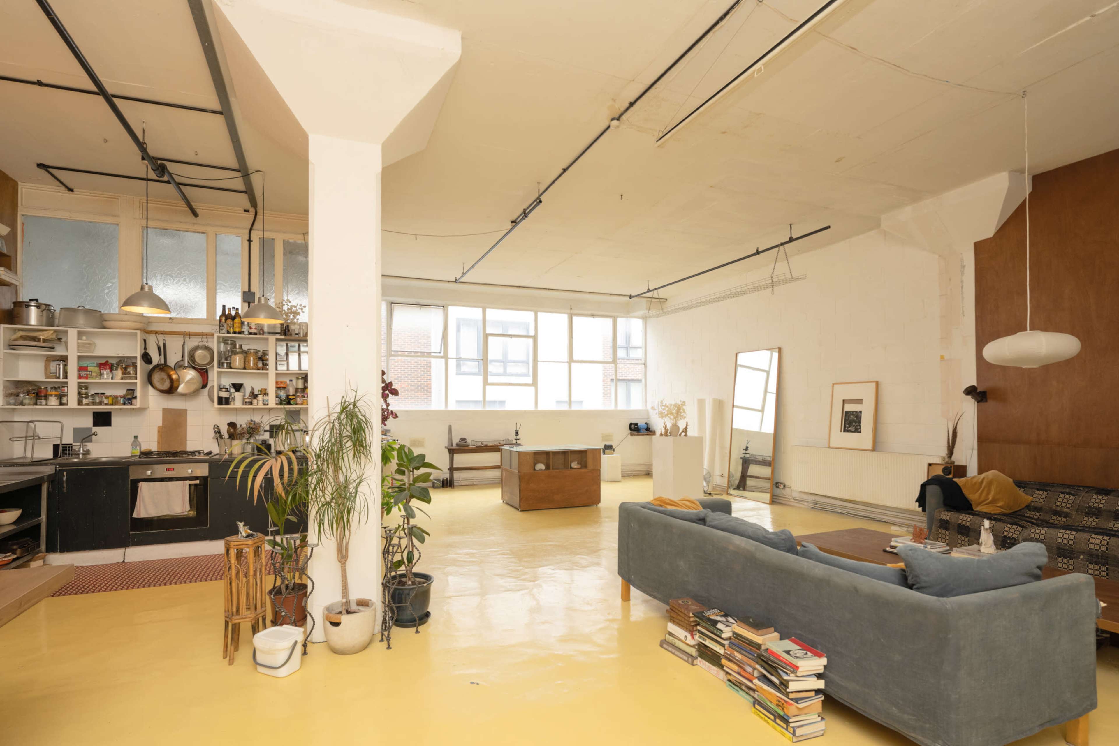 The image shows a spacious, brightly lit open-plan living area with a kitchen on one side, a sitting area with a couch and books, and large windows allowing natural light to flow in.