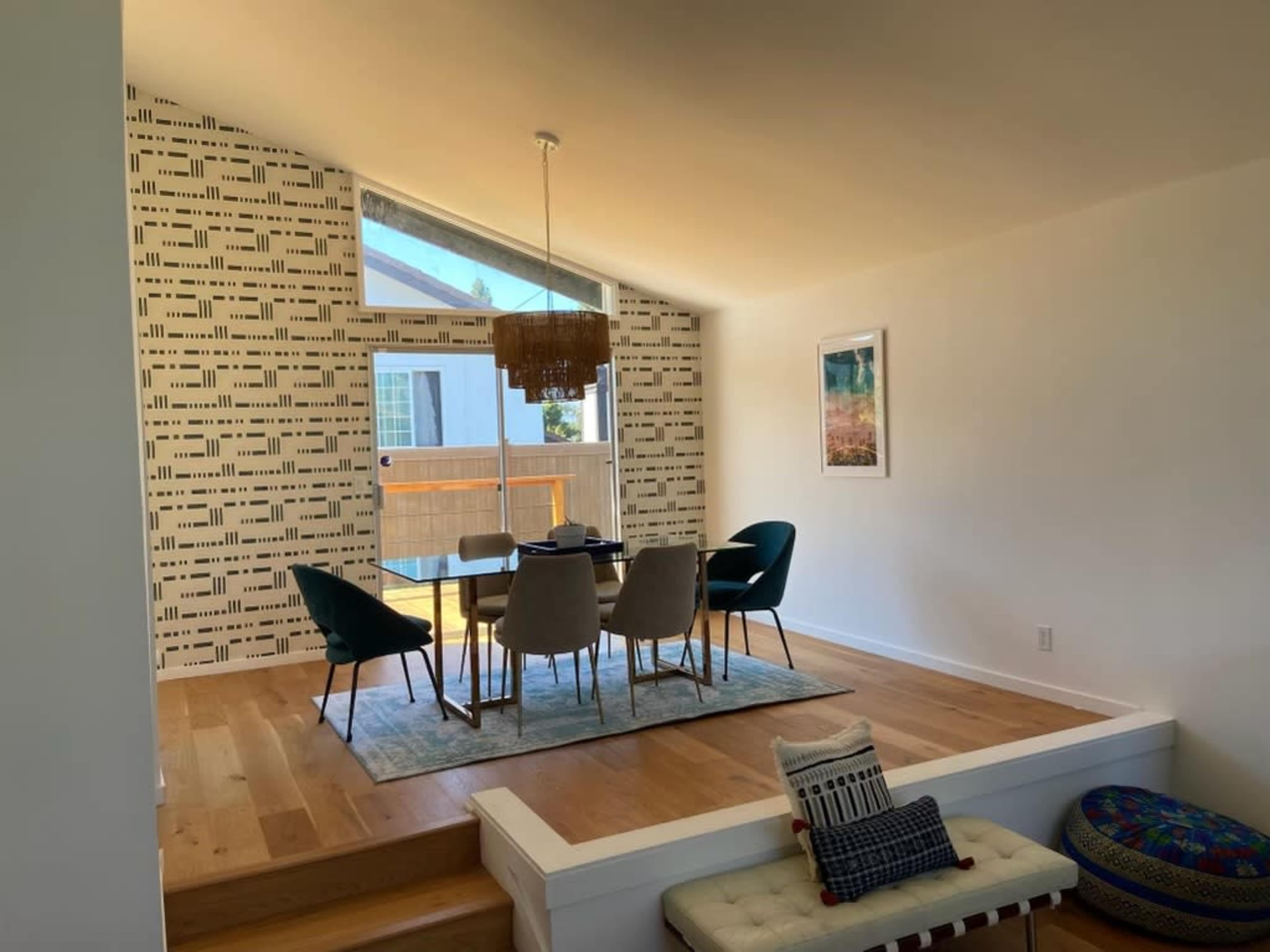 A dining area features a table surrounded by chairs and a large window with a patterned wall in the background.