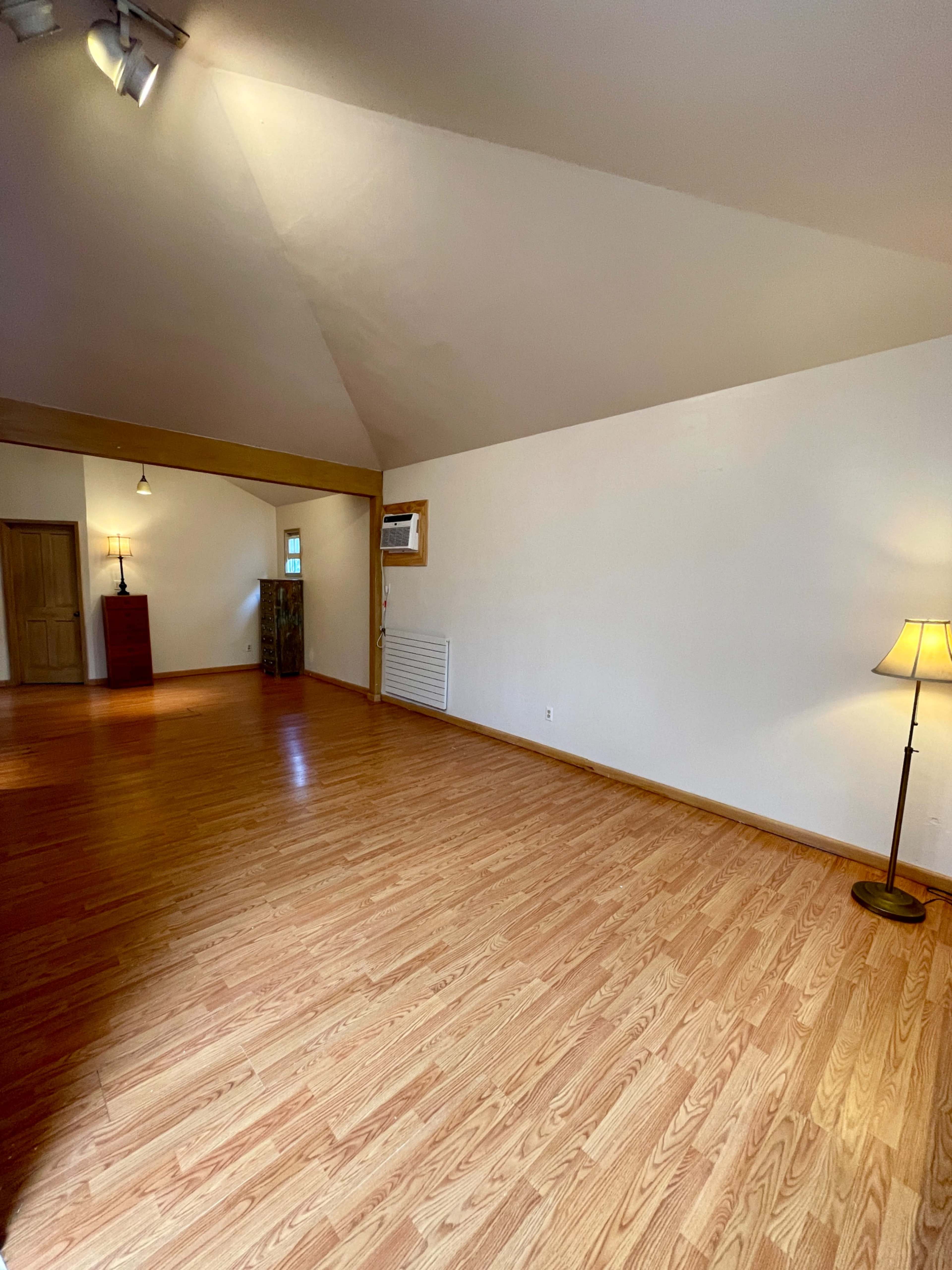 The image shows a spacious, empty room with hardwood flooring, a sloped ceiling, and a lamp in one corner.