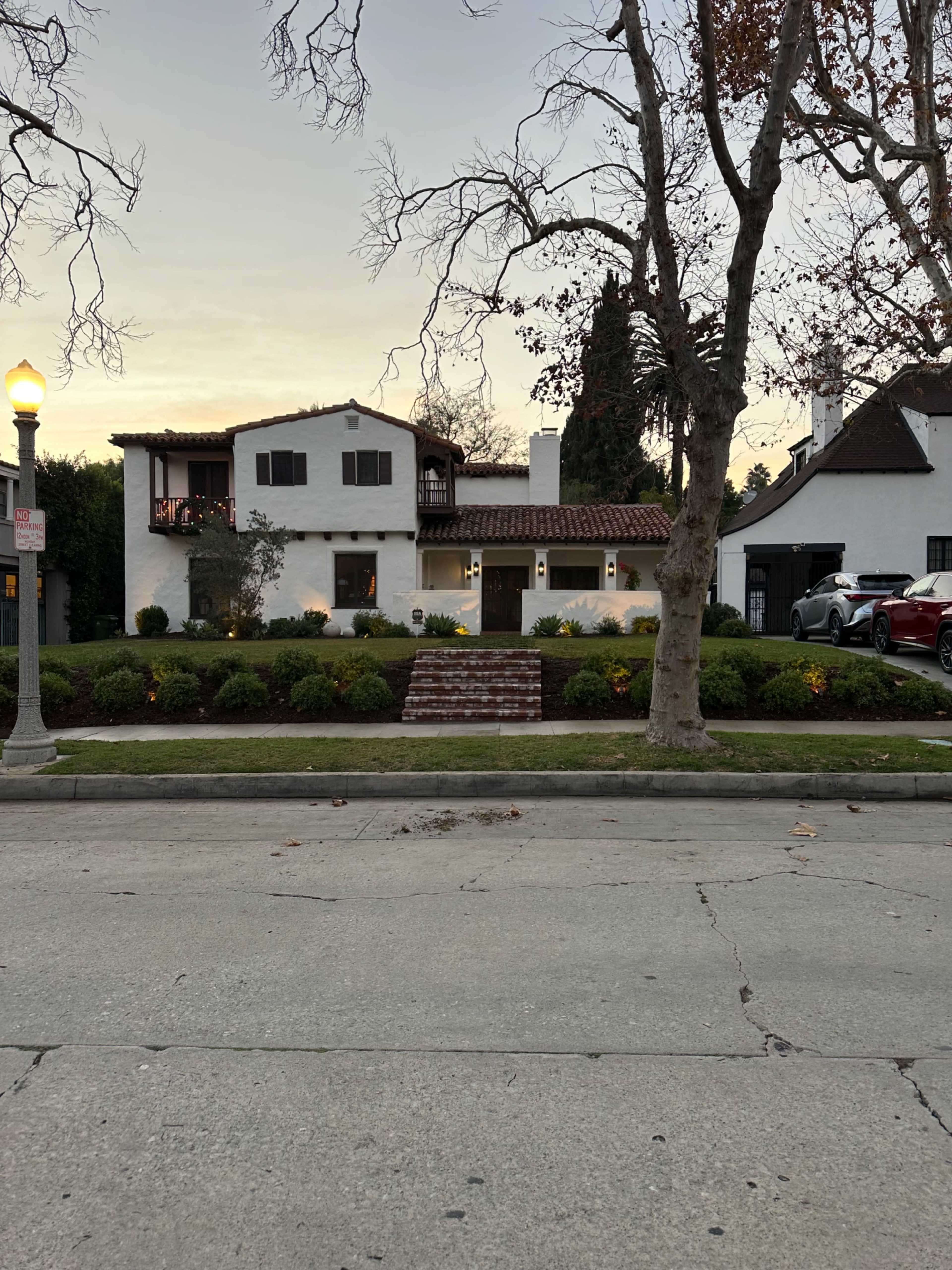 A white stucco house with a tiled roof and a front garden is situated along a street lined with trees.