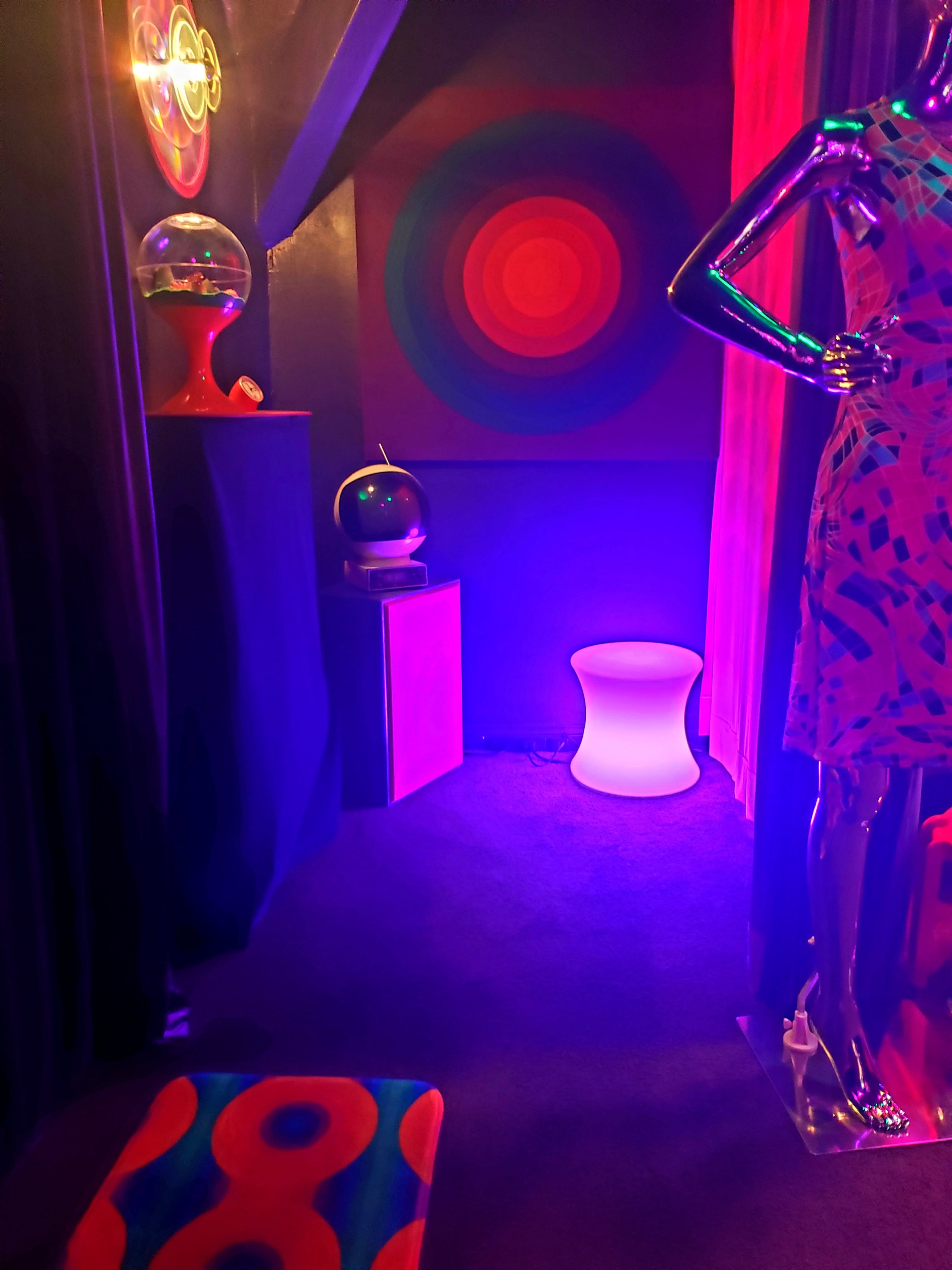 A dimly lit room with colorful decor, featuring a mannequin dressed in a vibrant outfit, a glowing stool, and a spiral-patterned wall.