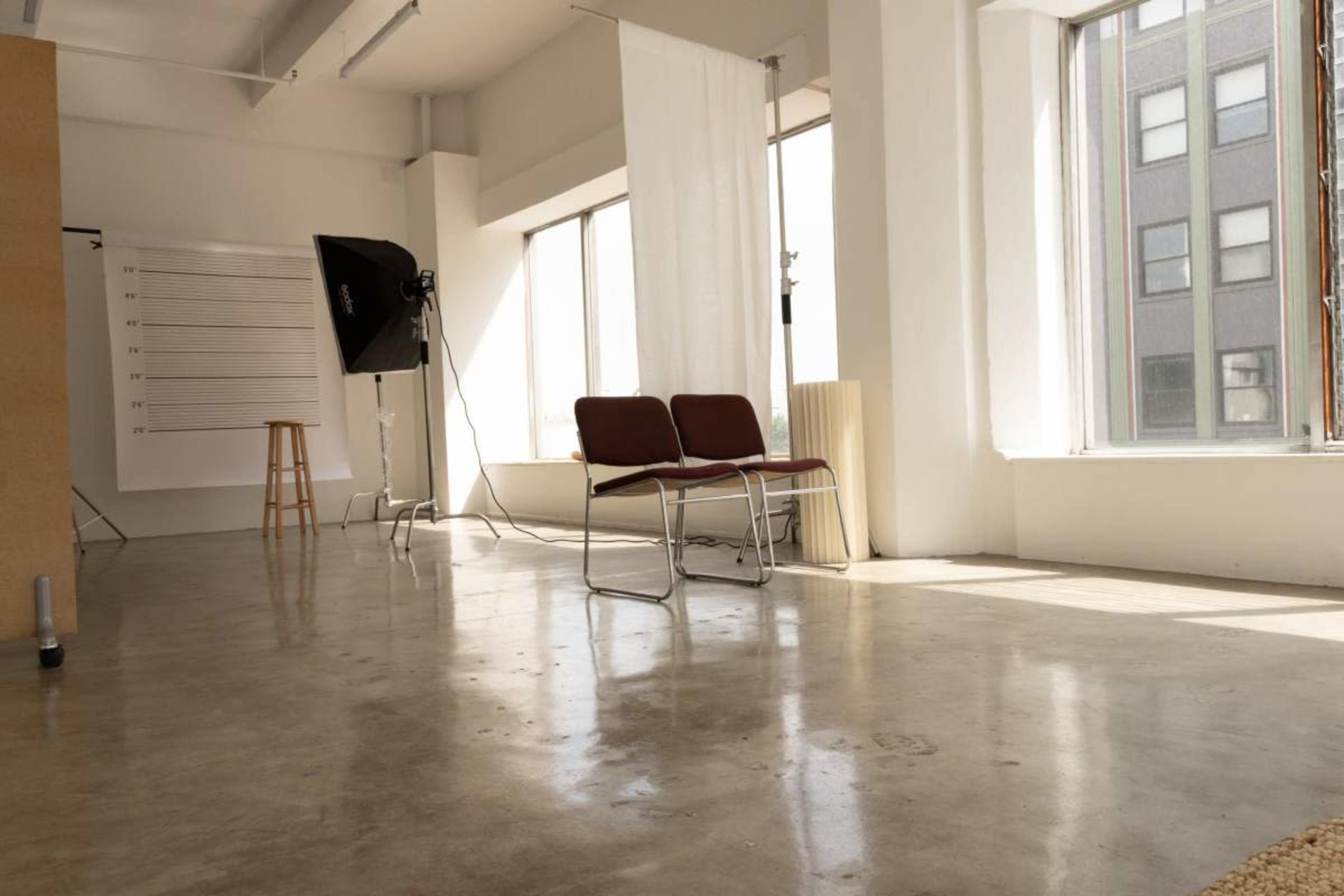 The image shows a spacious interior featuring two chairs facing a large window, with a backdrop and lighting equipment set up nearby.