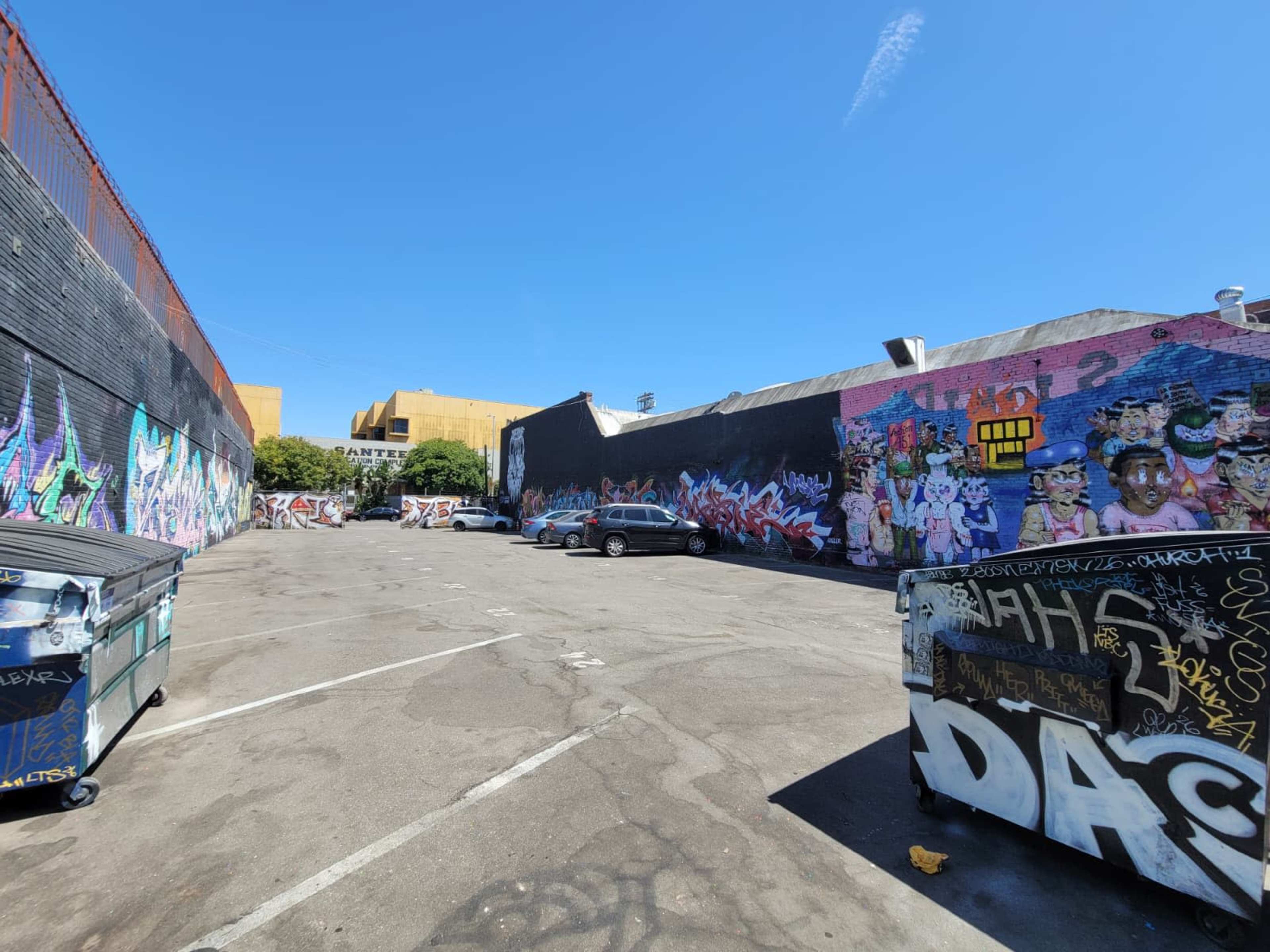 The image shows a narrow urban alleyway with colorful graffiti on the walls and several parked cars along the pavement.