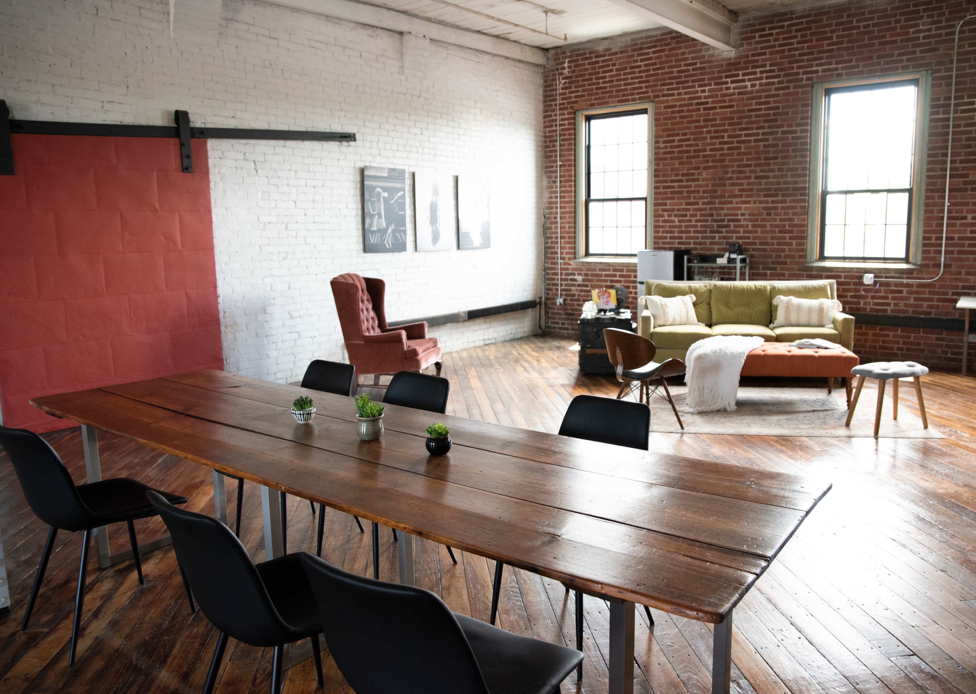 A spacious industrial room features a large wooden table with black chairs, a red accent wall, and a cozy seating area with a sofa and armchair.