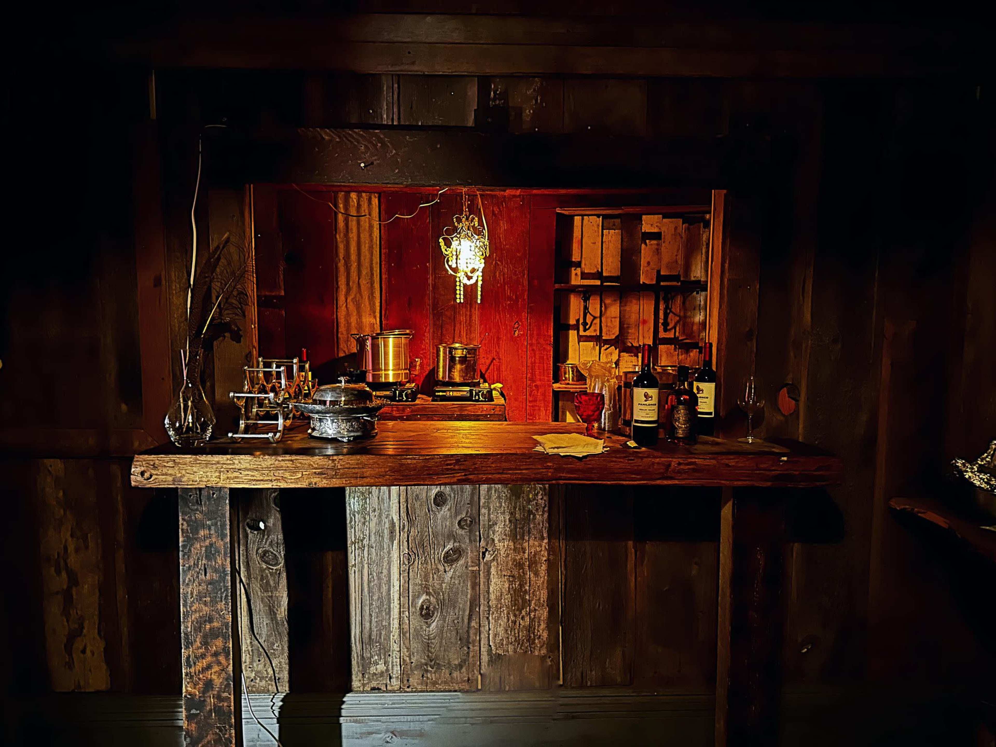 A wooden bar area with a small warm light overhead, showcasing pots and jars in the background against a rustic wall.