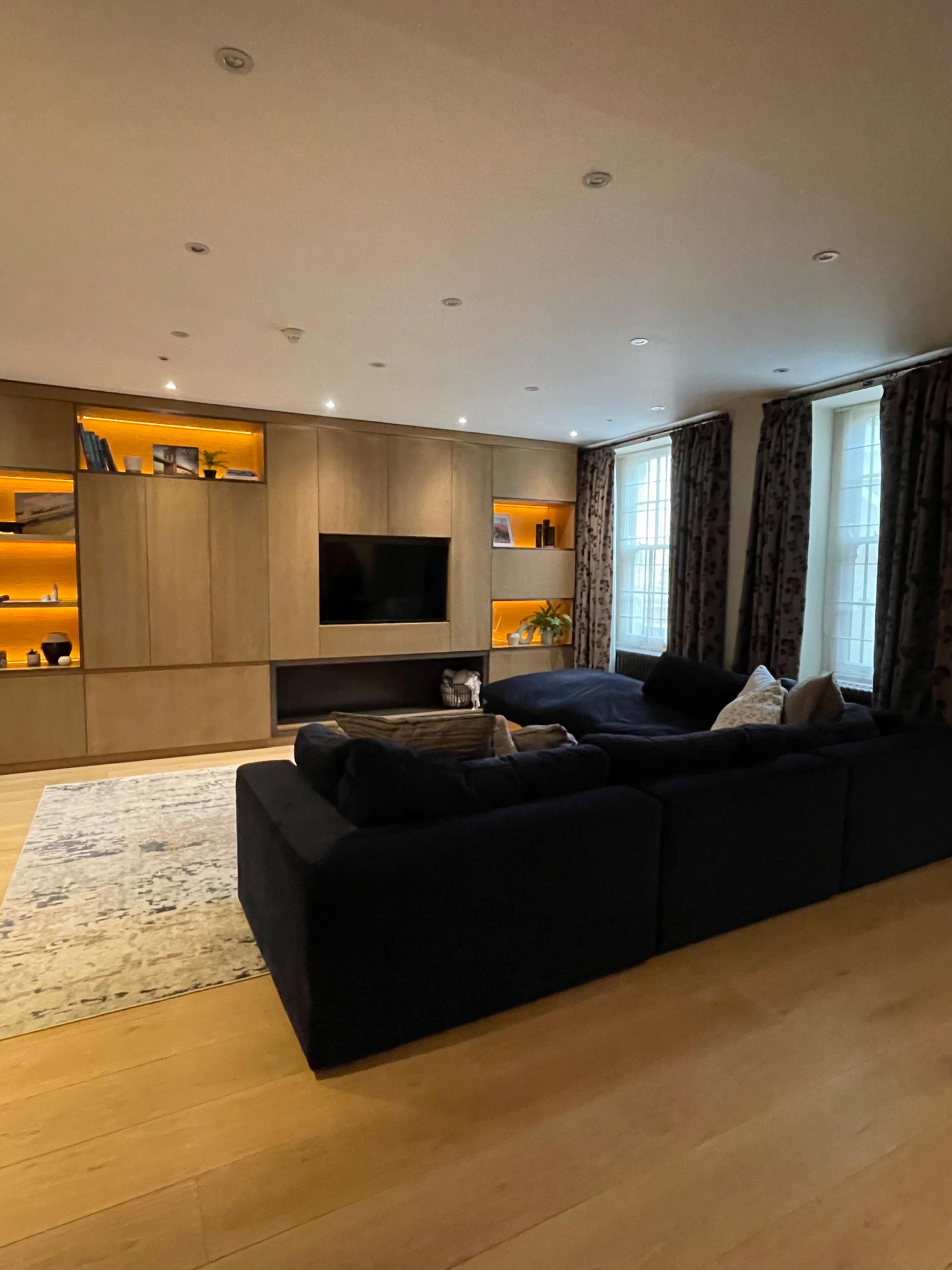 The image shows a modern living room with wooden cabinetry, a flat-screen TV, and a dark sectional sofa, complemented by illuminated shelves and large windows.