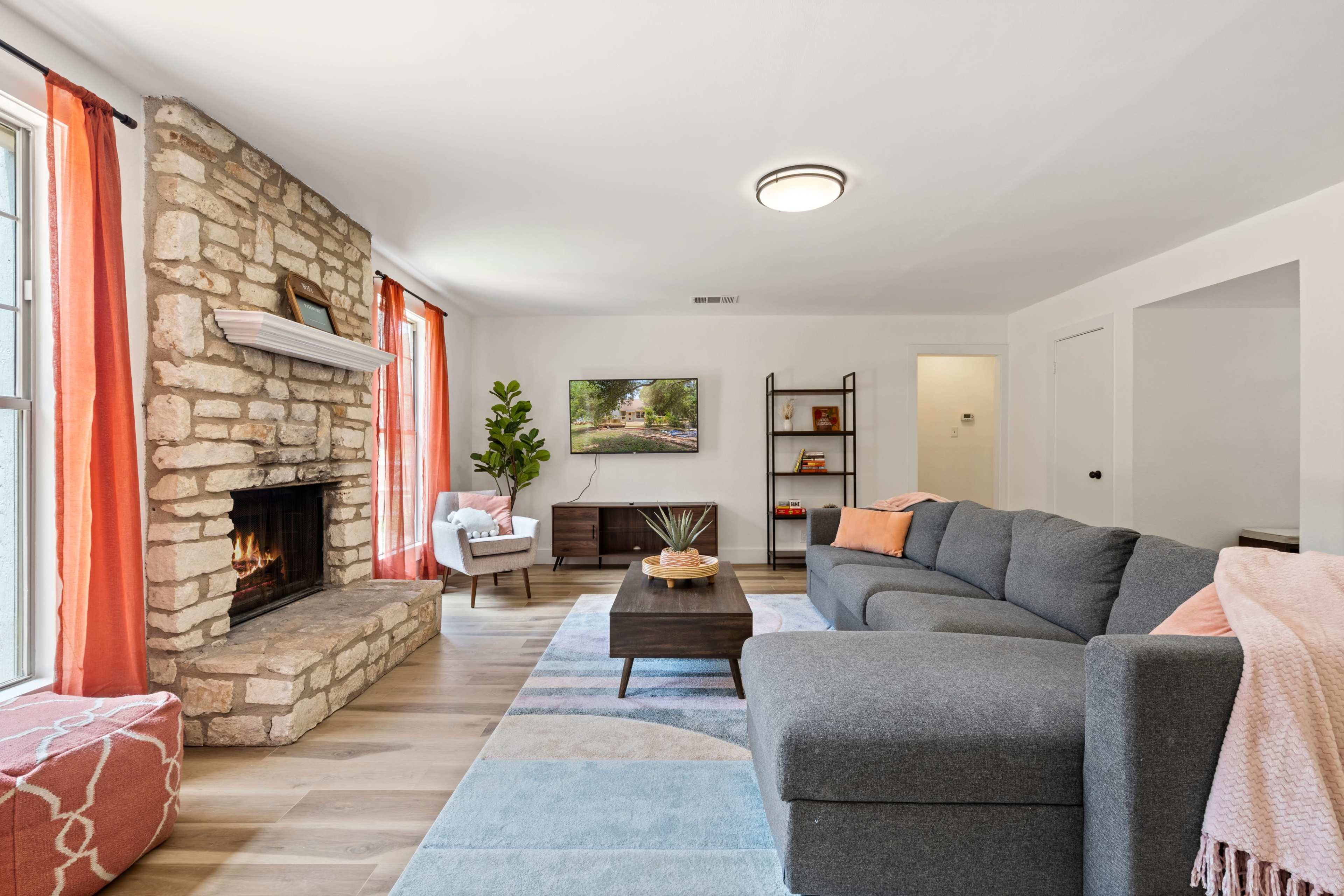 A cozy living room features a stone fireplace, modern furniture, and large windows with orange curtains.