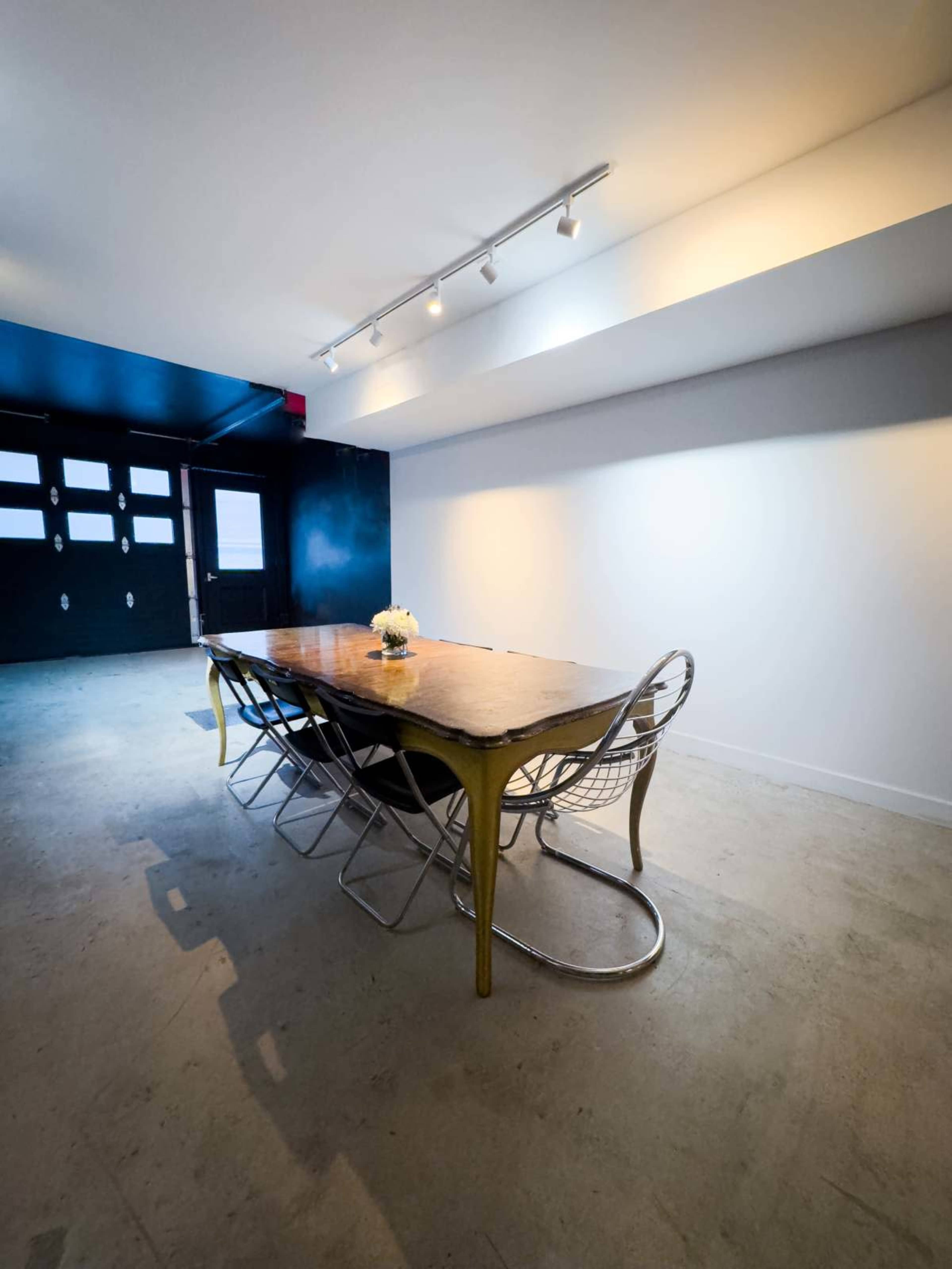 A long wooden dining table with metal chairs is set in a minimalist room featuring a black wall and large windows.