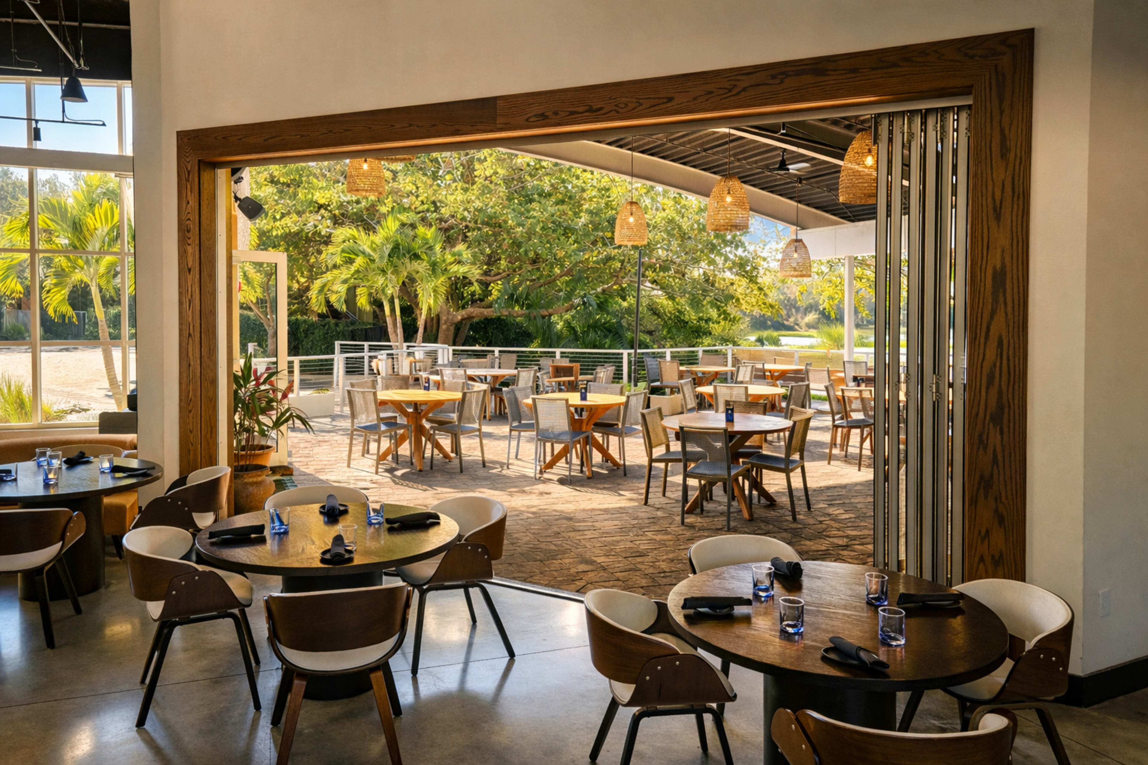 The image shows the interior of a restaurant with wooden tables and chairs, overlooking an outdoor dining area surrounded by greenery.
