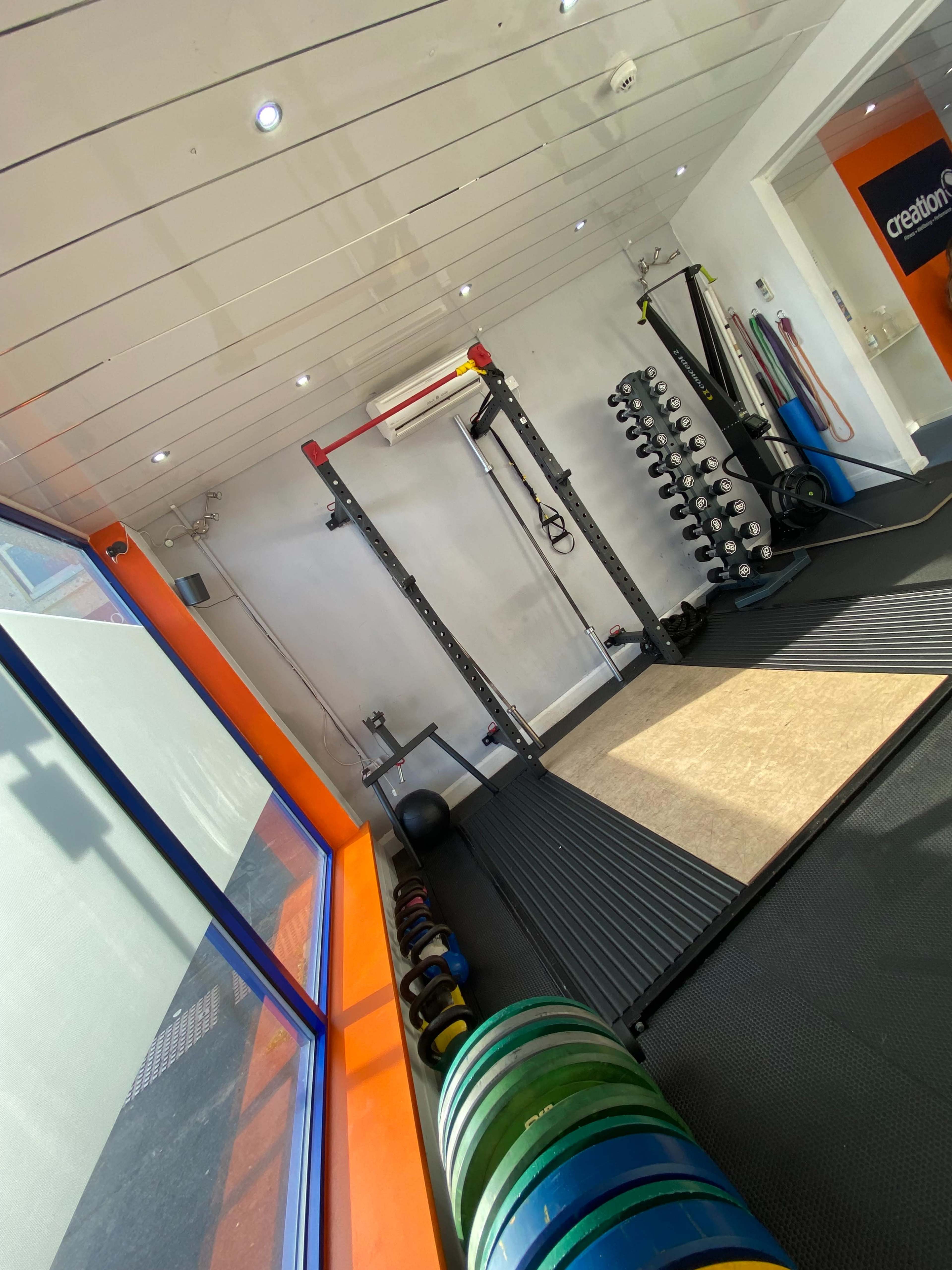 The image shows a well-lit gym space featuring weightlifting equipment, a squat rack, dumbbells, and exercise mats.