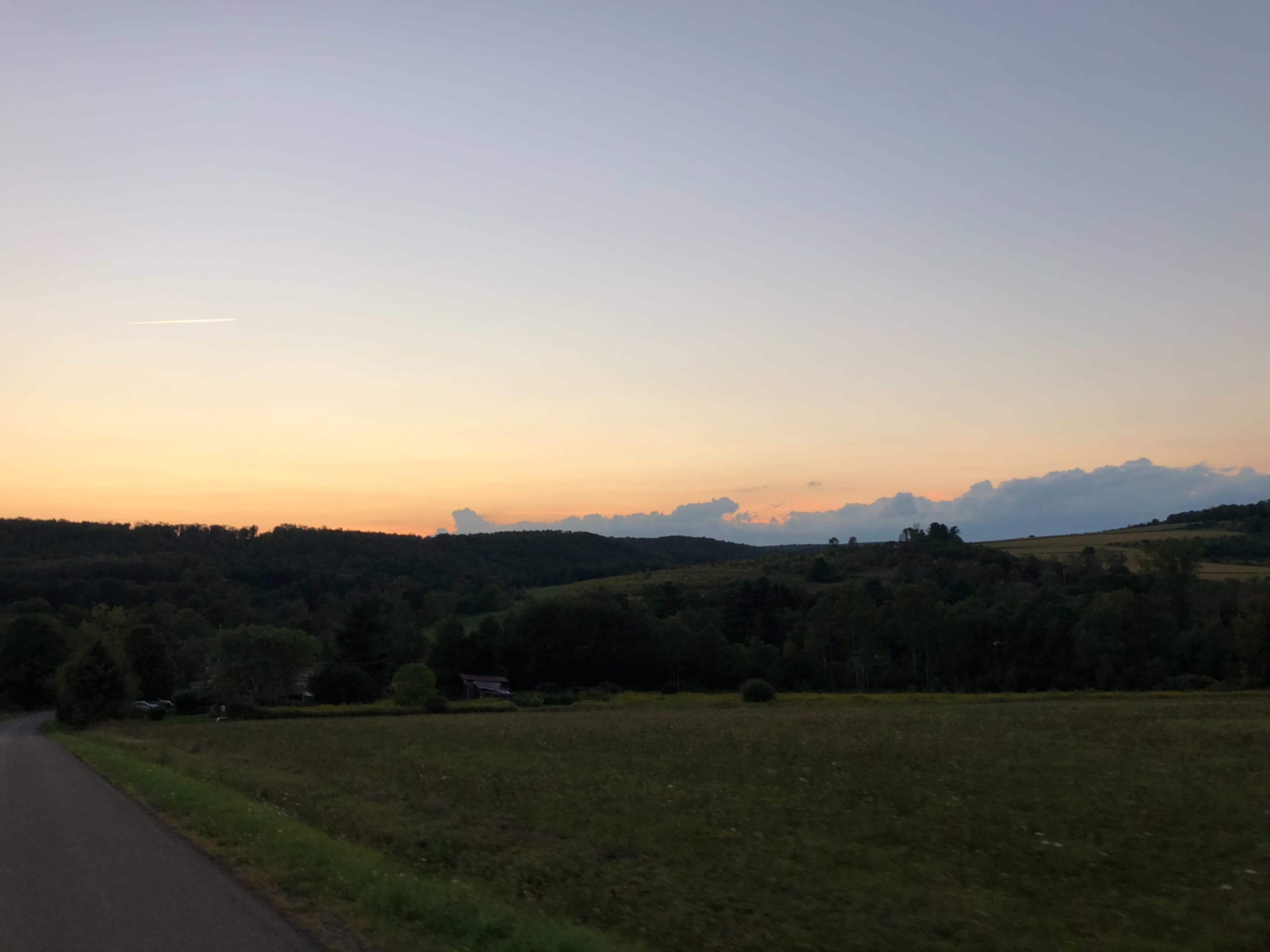 A paved road leads through a grassy field toward a horizon of rolling hills under a gradient sky at dusk.
