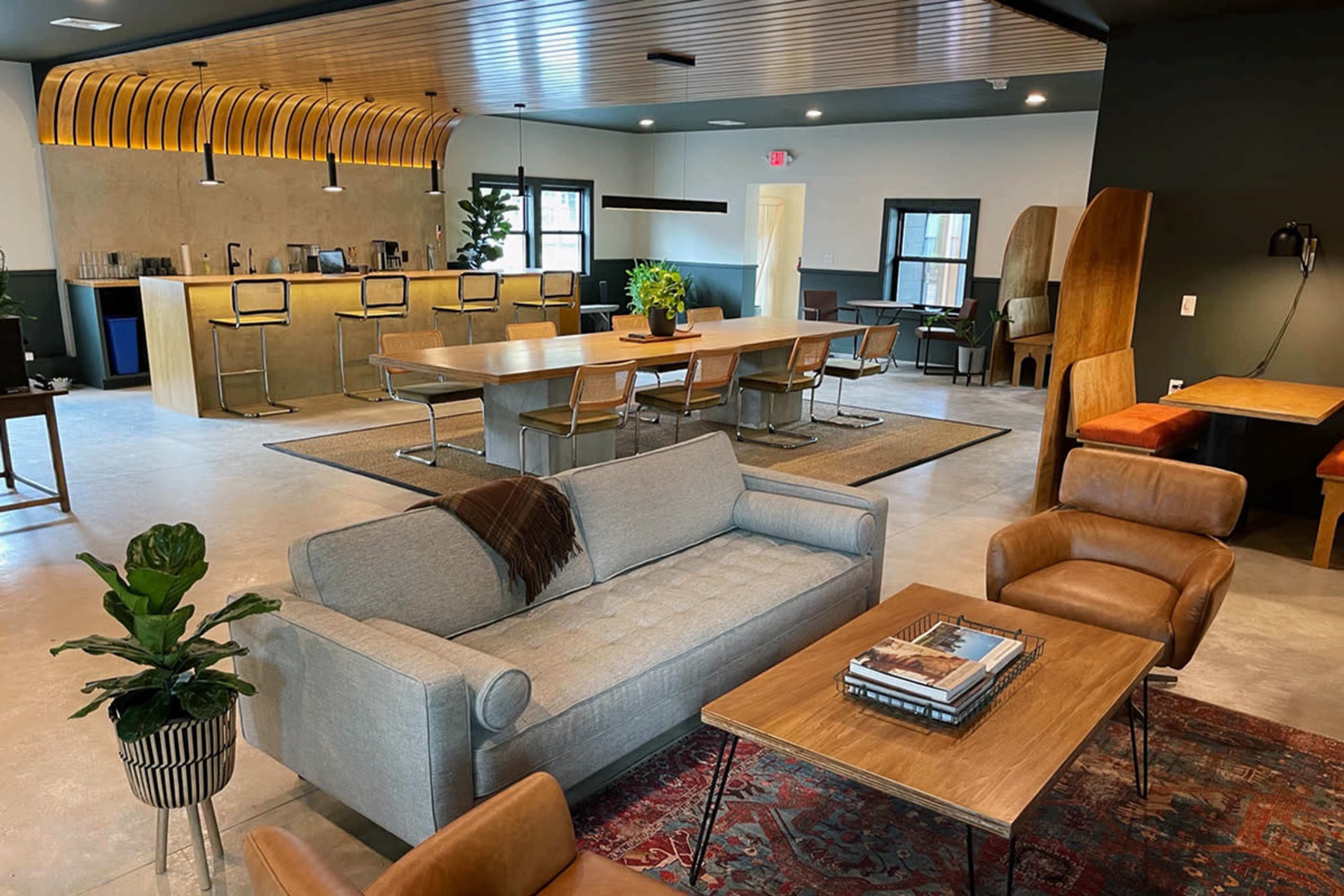 A modern interior space featuring a communal seating area with a sofa and armchairs, a large wooden dining table, and a bar counter in the background.