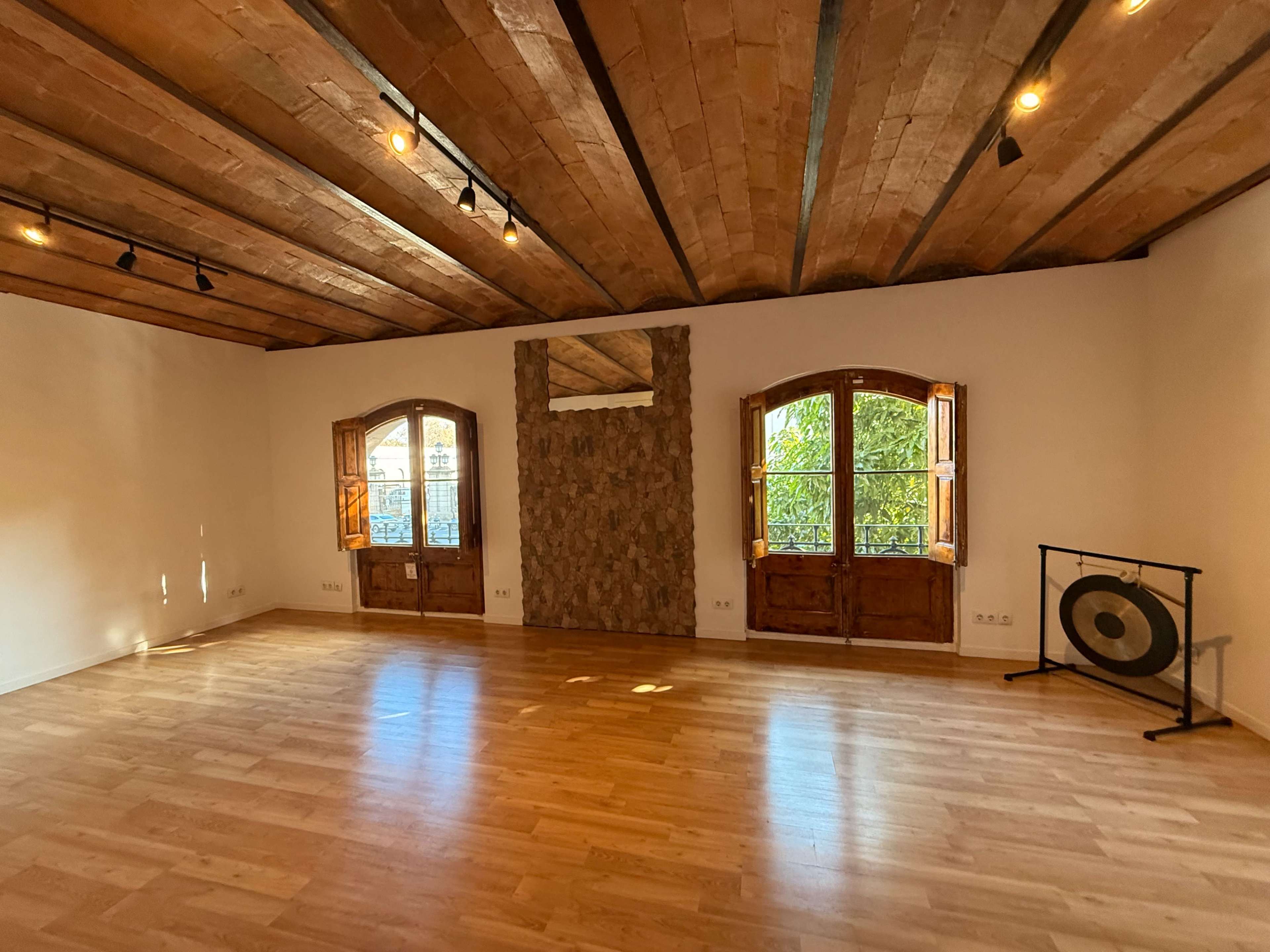 The image shows a spacious room with wooden beams on the ceiling, two large windows, a stone feature wall, and a gong on a stand in one corner.