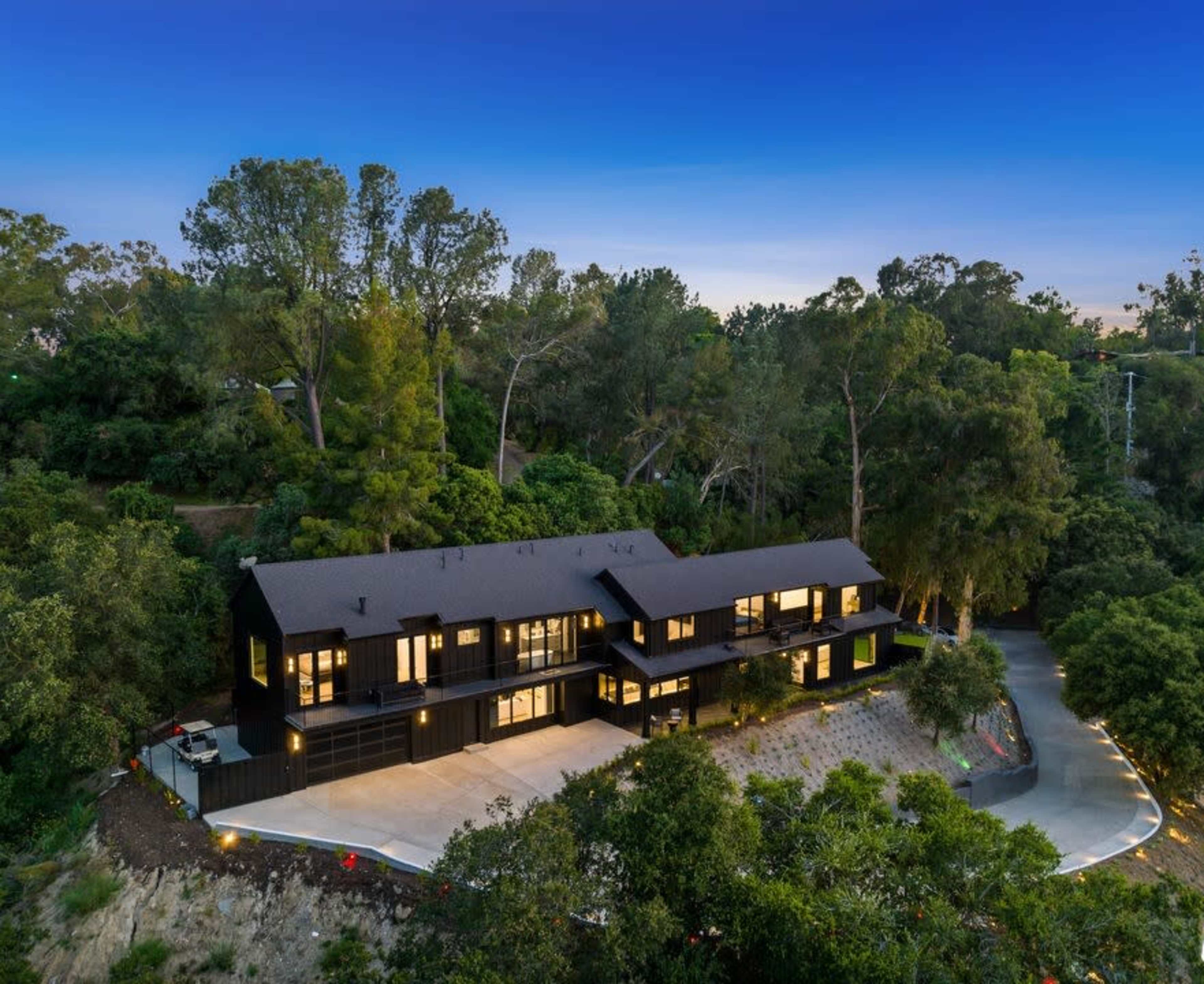 A modern black house is situated on a hillside, surrounded by trees, with a winding driveway leading to it.