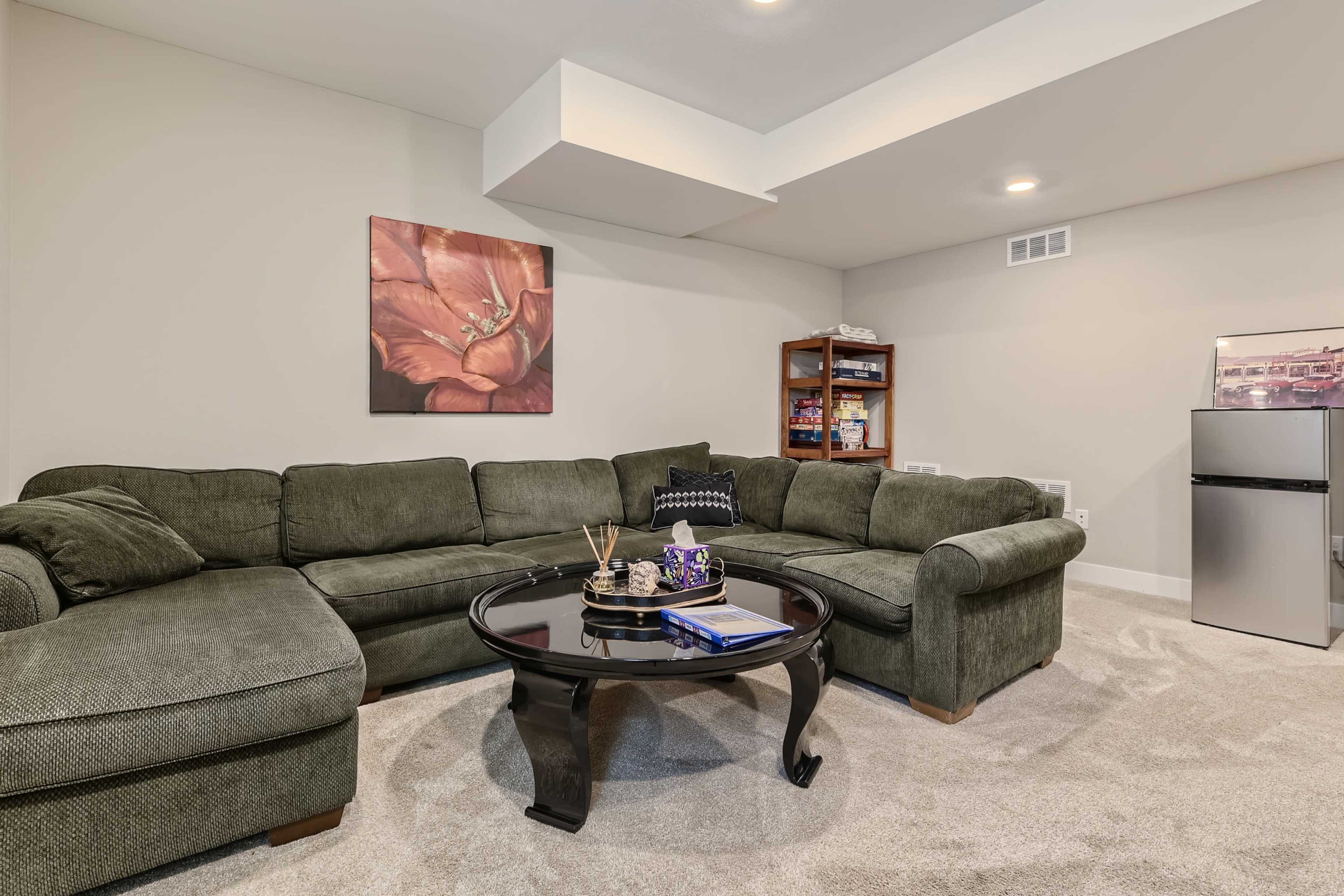A spacious living area features a large sectional couch, a round coffee table, and a small refrigerator, with a floral artwork hanging on the wall.