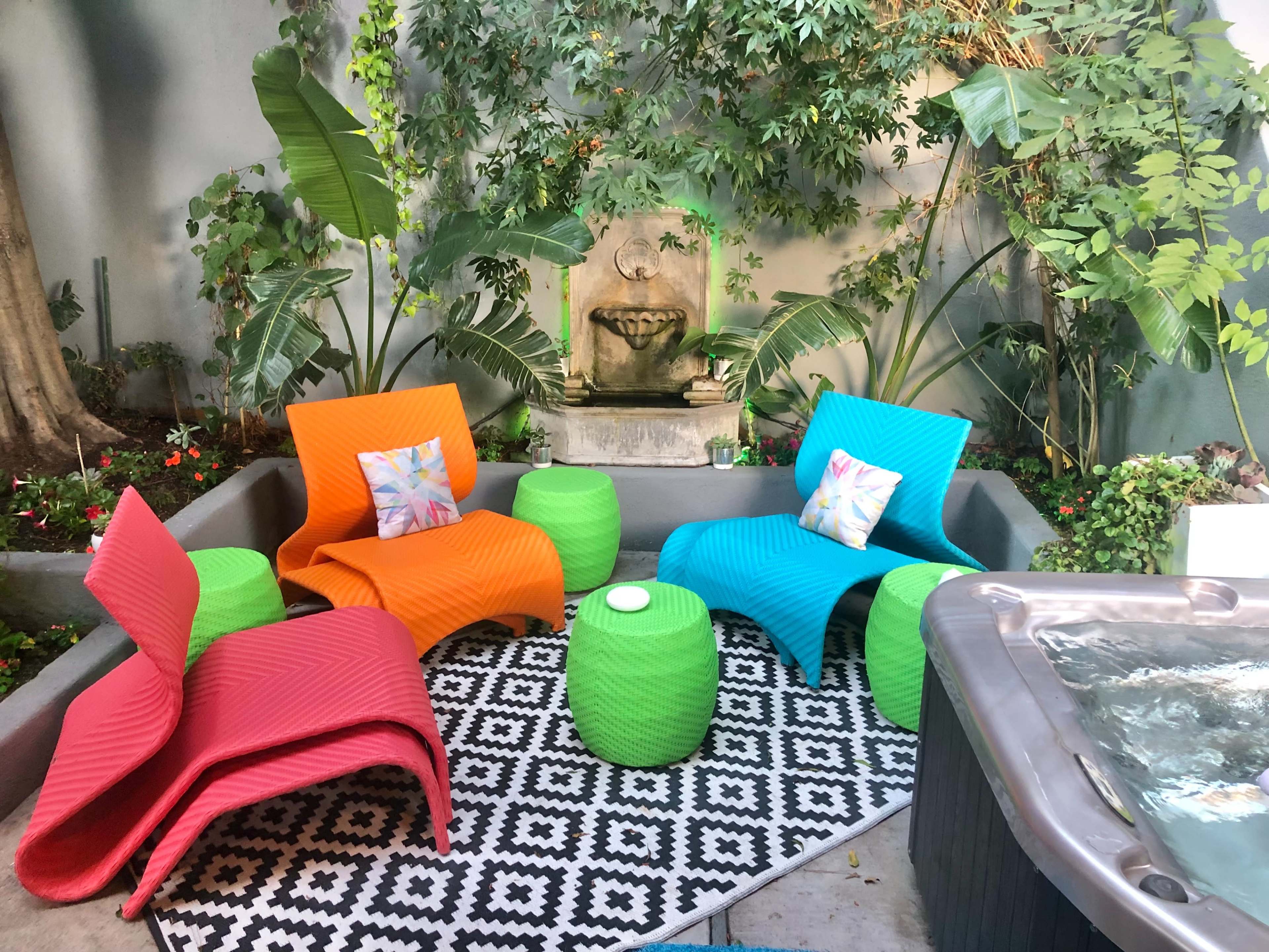 The image shows a vibrant outdoor seating area with colorful chairs, a patterned rug, potted plants, and a small fountain.