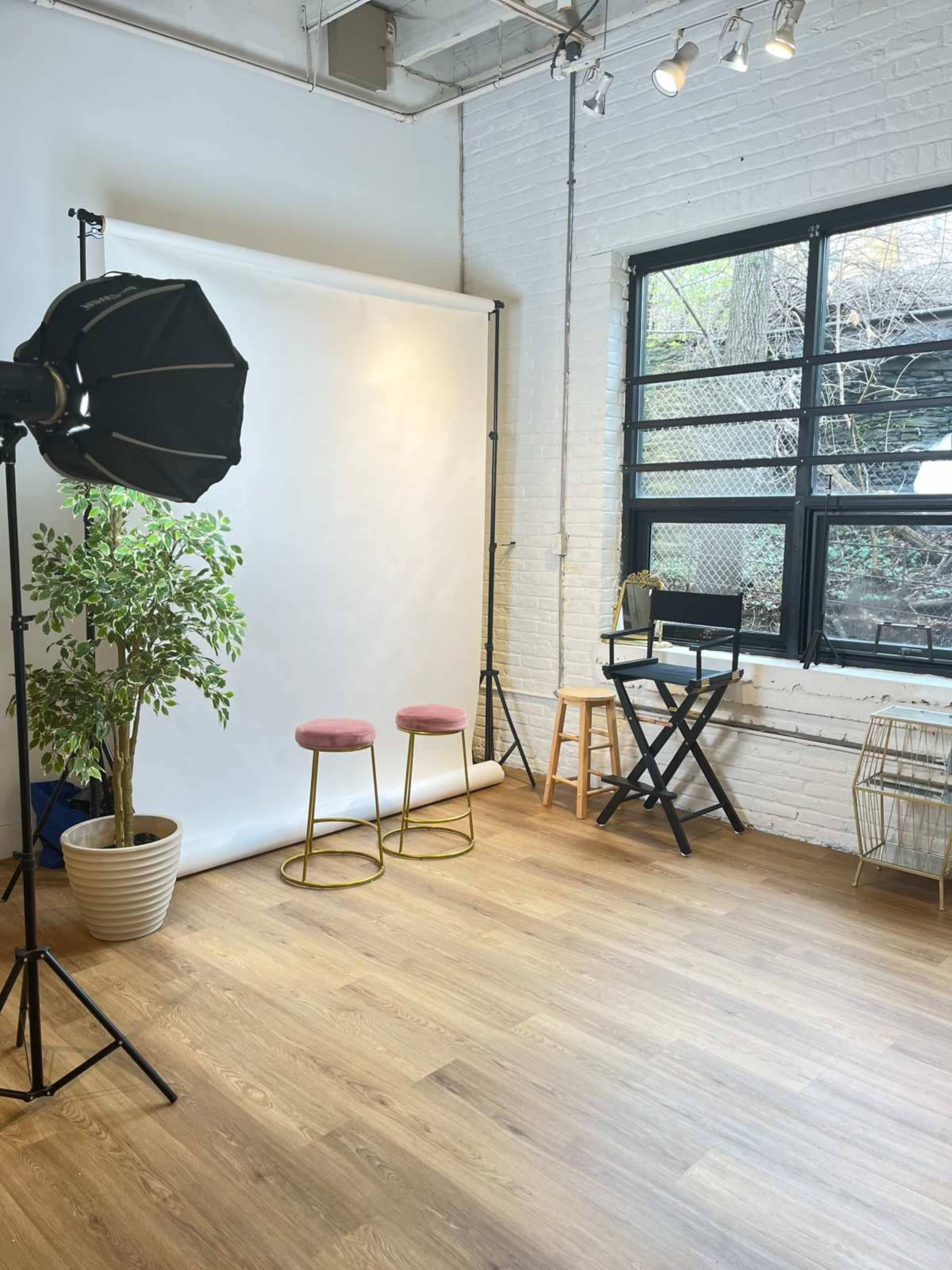 A photography studio with a white backdrop, two chairs, a plant, and a director's chair near a large window.