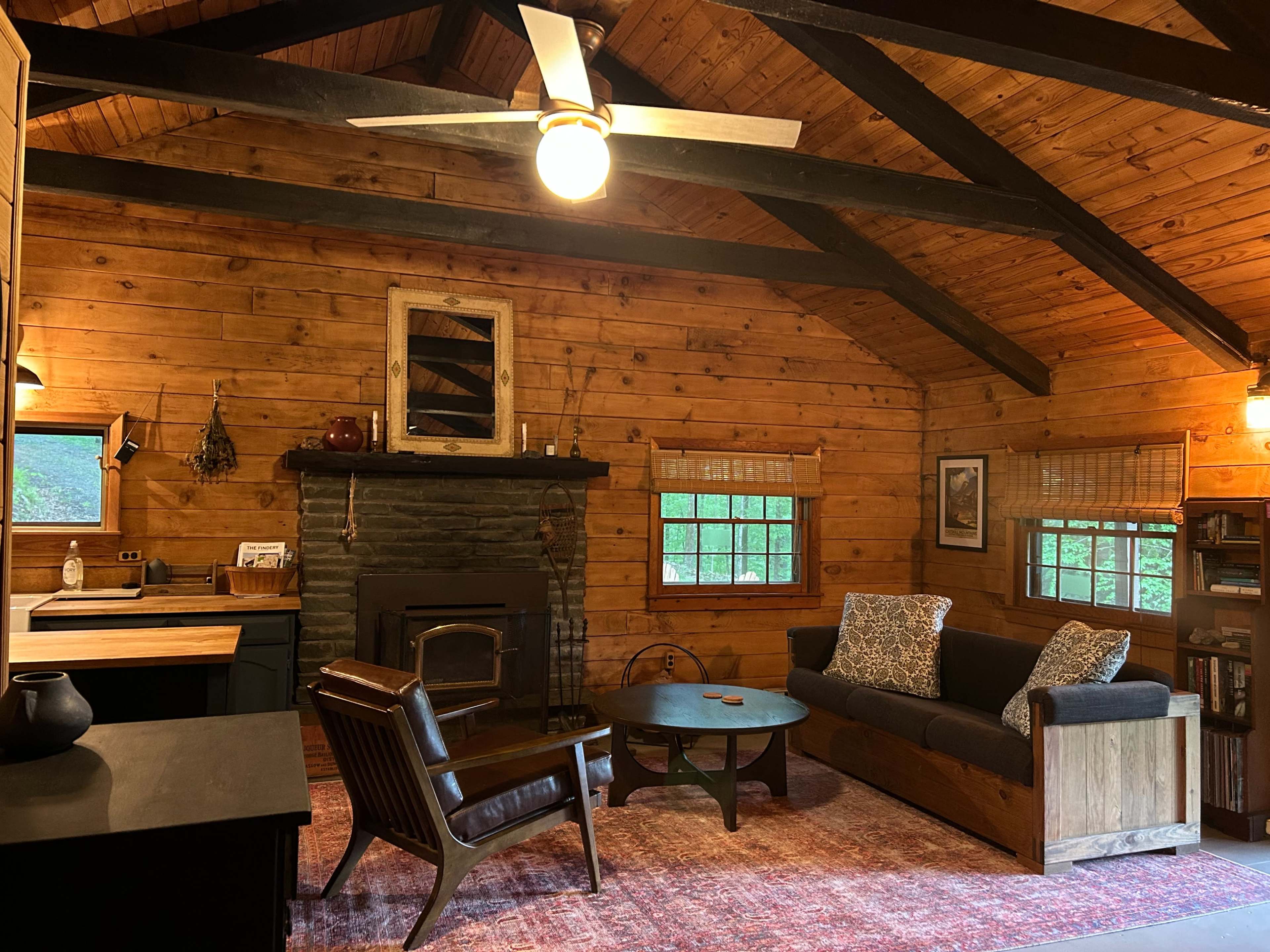 The image shows a rustic cabin interior with wood-paneled walls, a stone fireplace, a brown sofa, and a round coffee table set beneath exposed wooden beams.