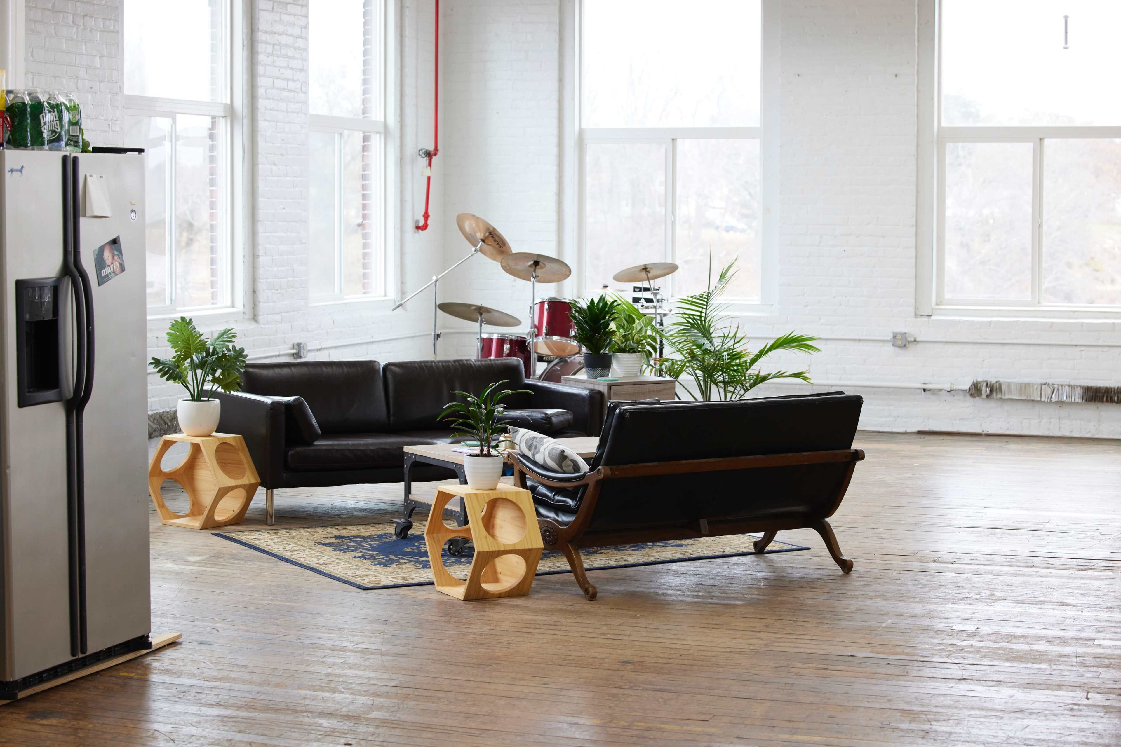 A spacious, well-lit room with two black sofas, a coffee table, a few potted plants, a modern-style rug, and a drum set in the corner.