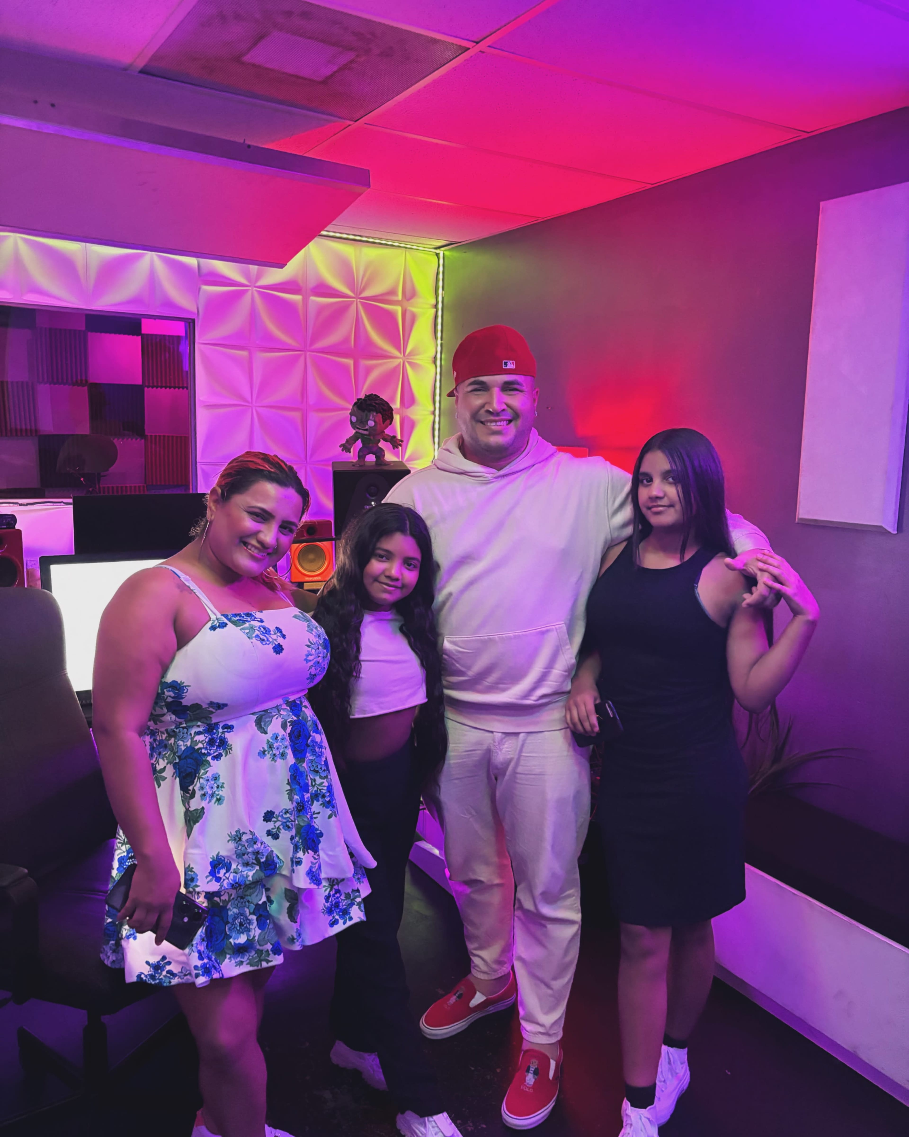 Four individuals are standing together in a brightly lit music studio, with colorful lighting and sound equipment visible in the background.