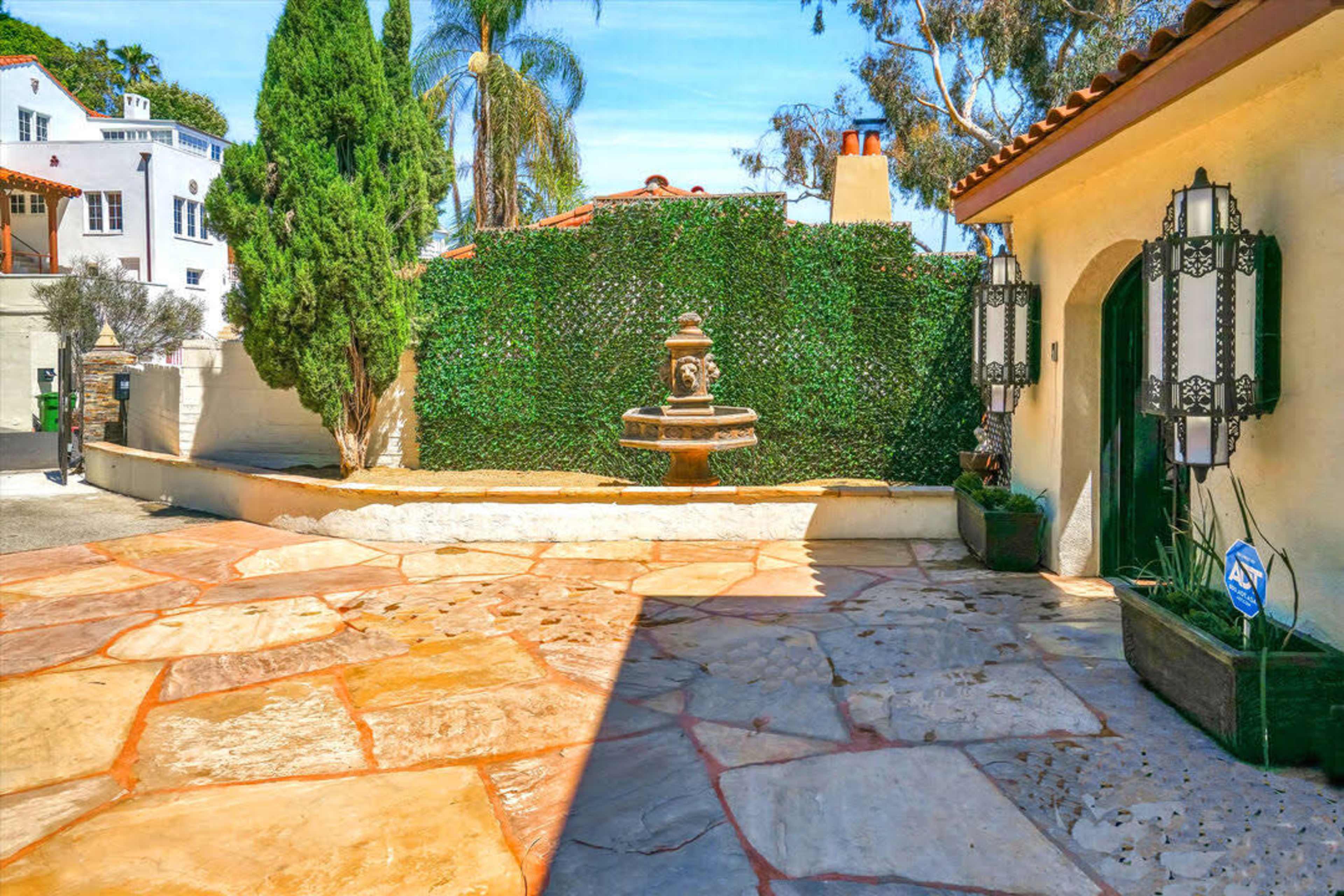 The image shows a stone-paved courtyard featuring a fountain surrounded by lush greenery and a building with a decorative doorway.