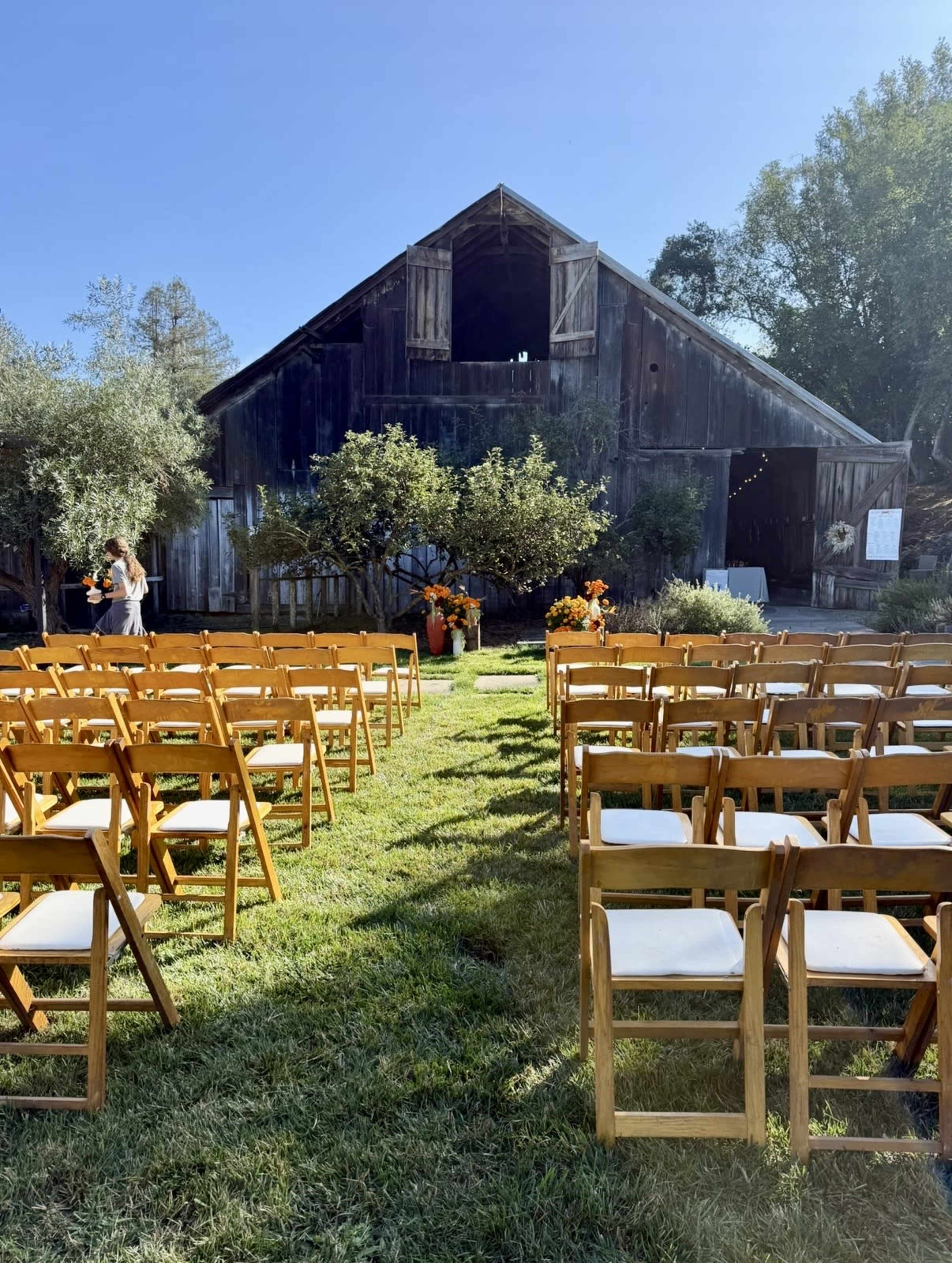 A rustic barn serves as the backdrop for a wedding ceremony, with rows of wooden chairs arranged on a grassy area.