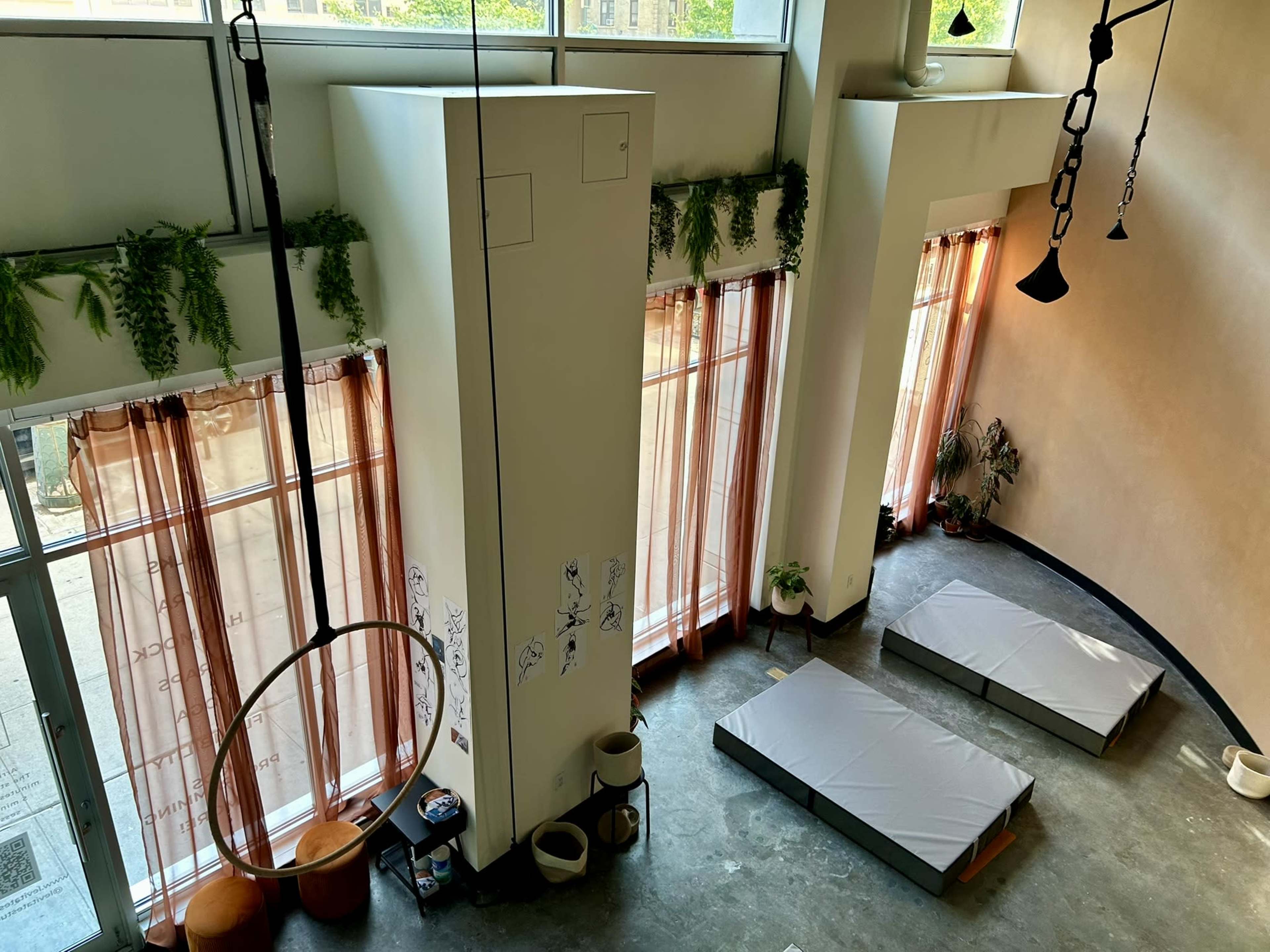 The image shows a spacious room with large windows, light curtains, and two gray mattresses laid out on the floor.