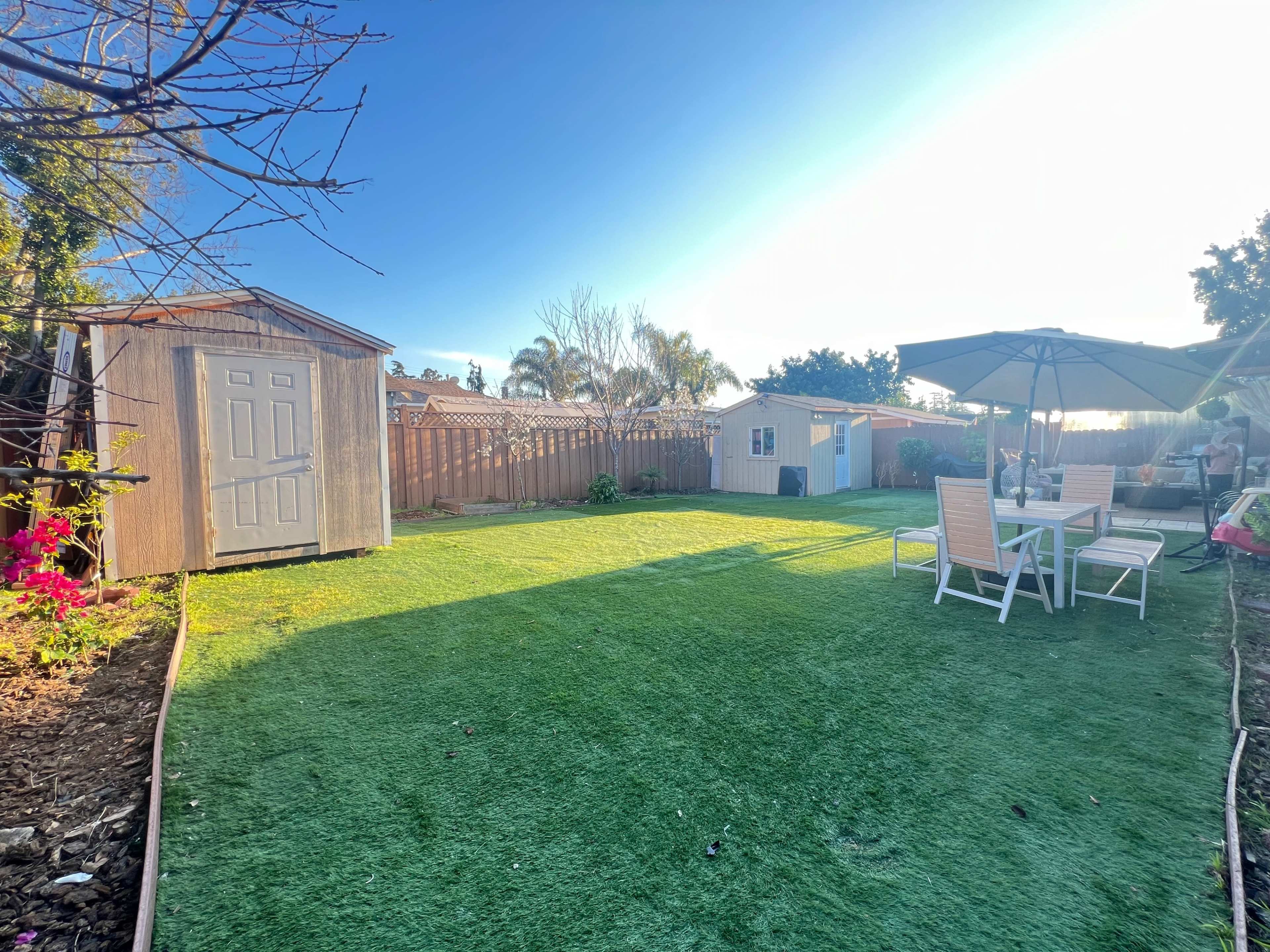 A backyard with artificial grass, a shed, a small white building, a patio umbrella, and outdoor furniture positioned on a sunny day.