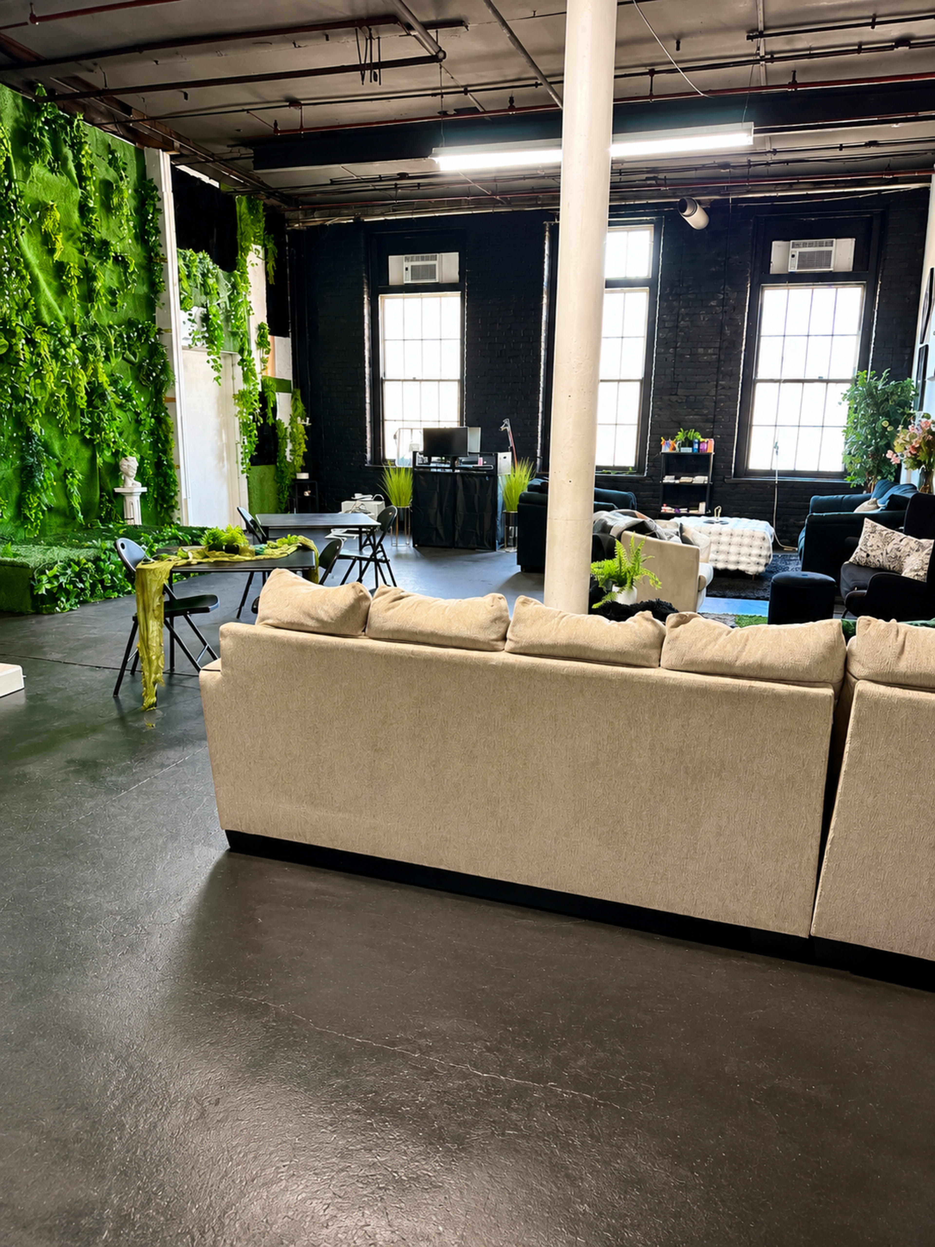 The image shows a spacious room featuring a beige sofa, green plants on the walls, and various seating arrangements on a dark floor.