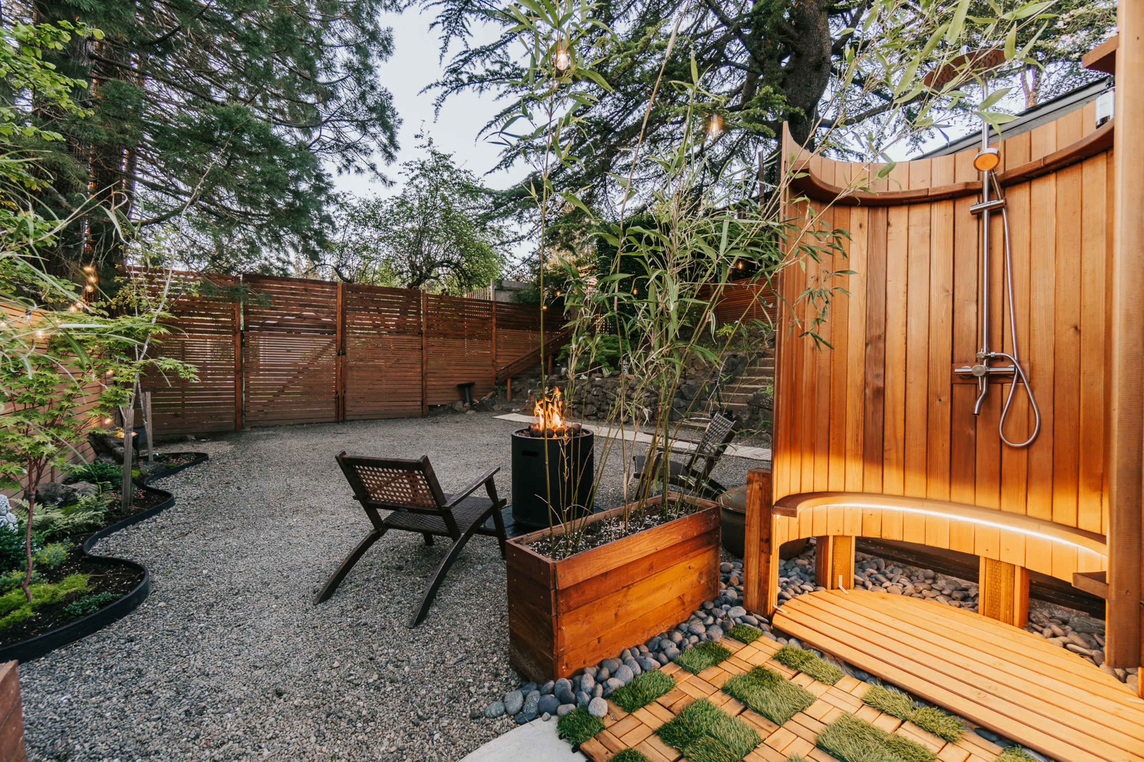 The image shows an outdoor shower area enclosed with wooden panels, featuring a wooden bench, a small fire pit, and landscaped gravel with plants.