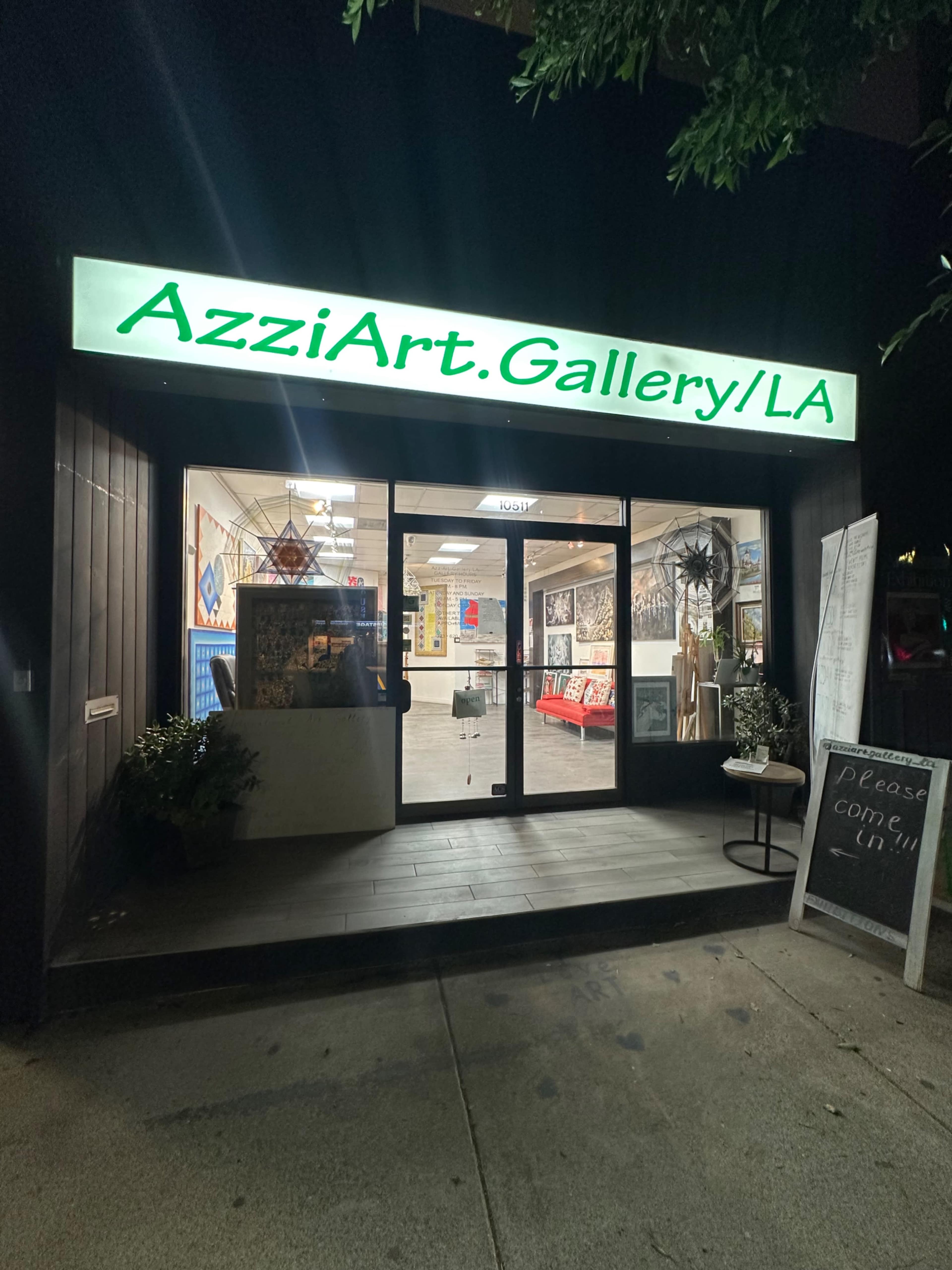 The exterior of AzziArt Gallery in LA is illuminated at night, showcasing its entrance with artwork displayed inside.
