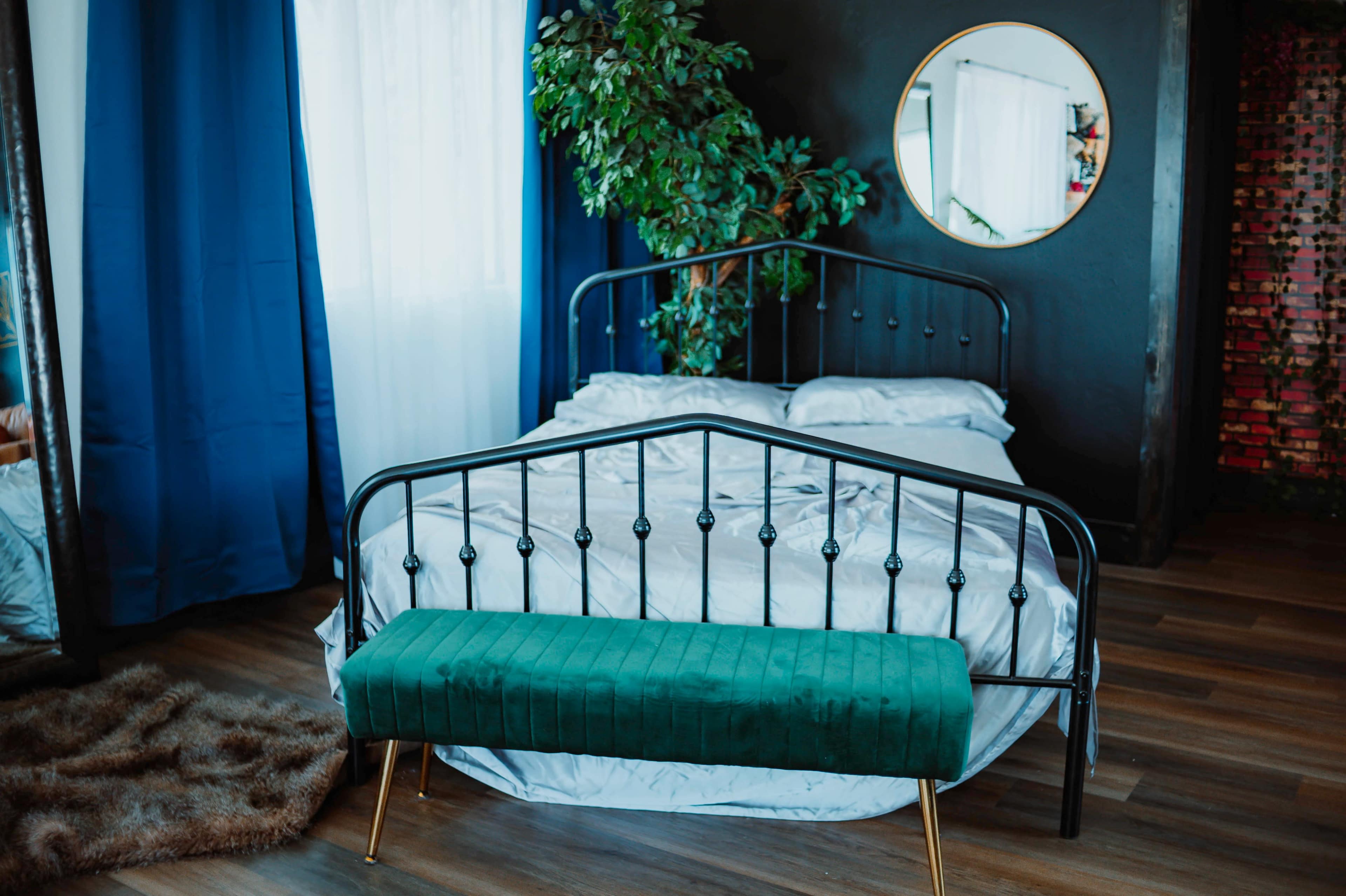 The image shows a modern bedroom featuring a black metal bed frame, a green bench at the foot of the bed, and large curtains beside a round mirror.