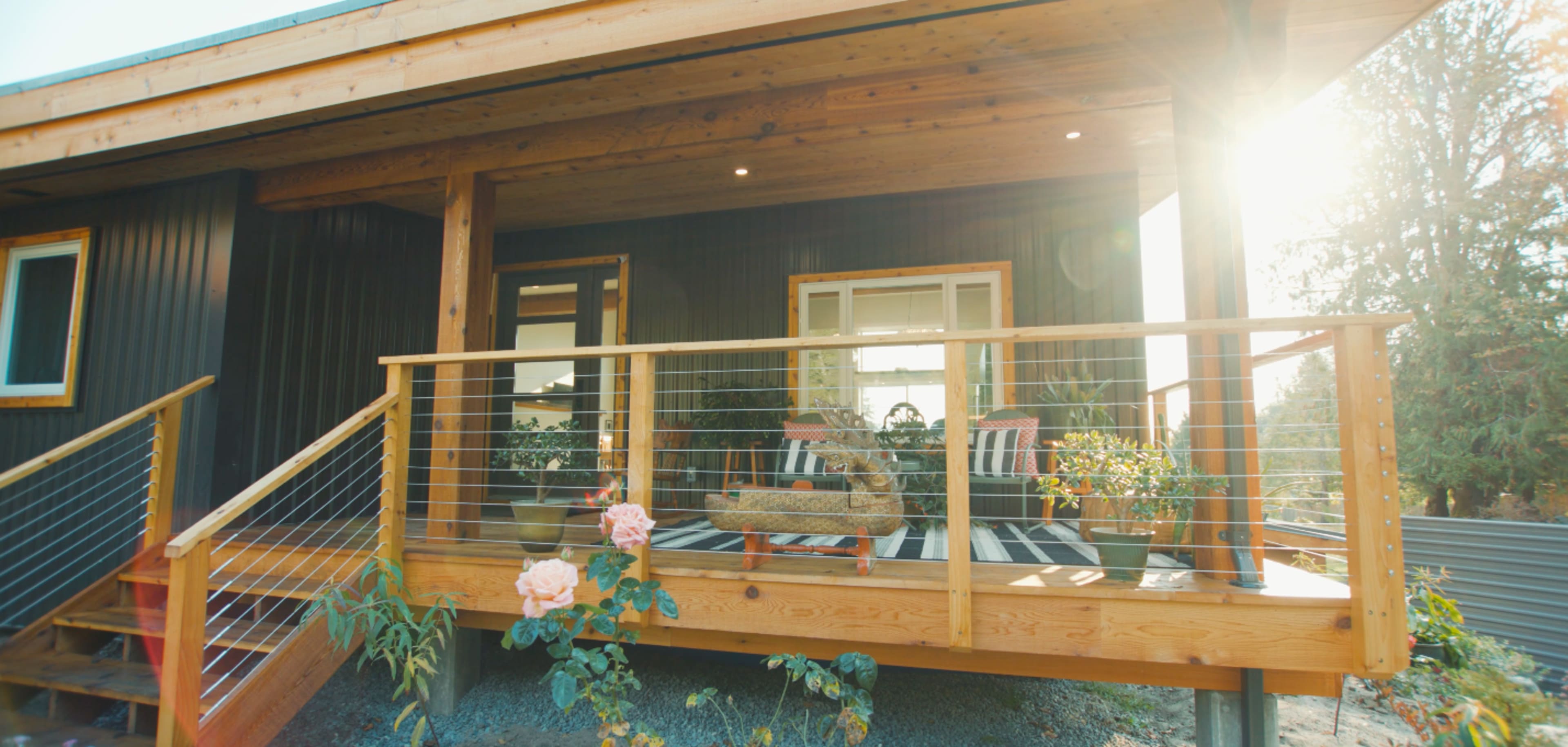 A front porch of a house, featuring a wooden deck with seating arrangements, potted plants, and sunlight streaming in.