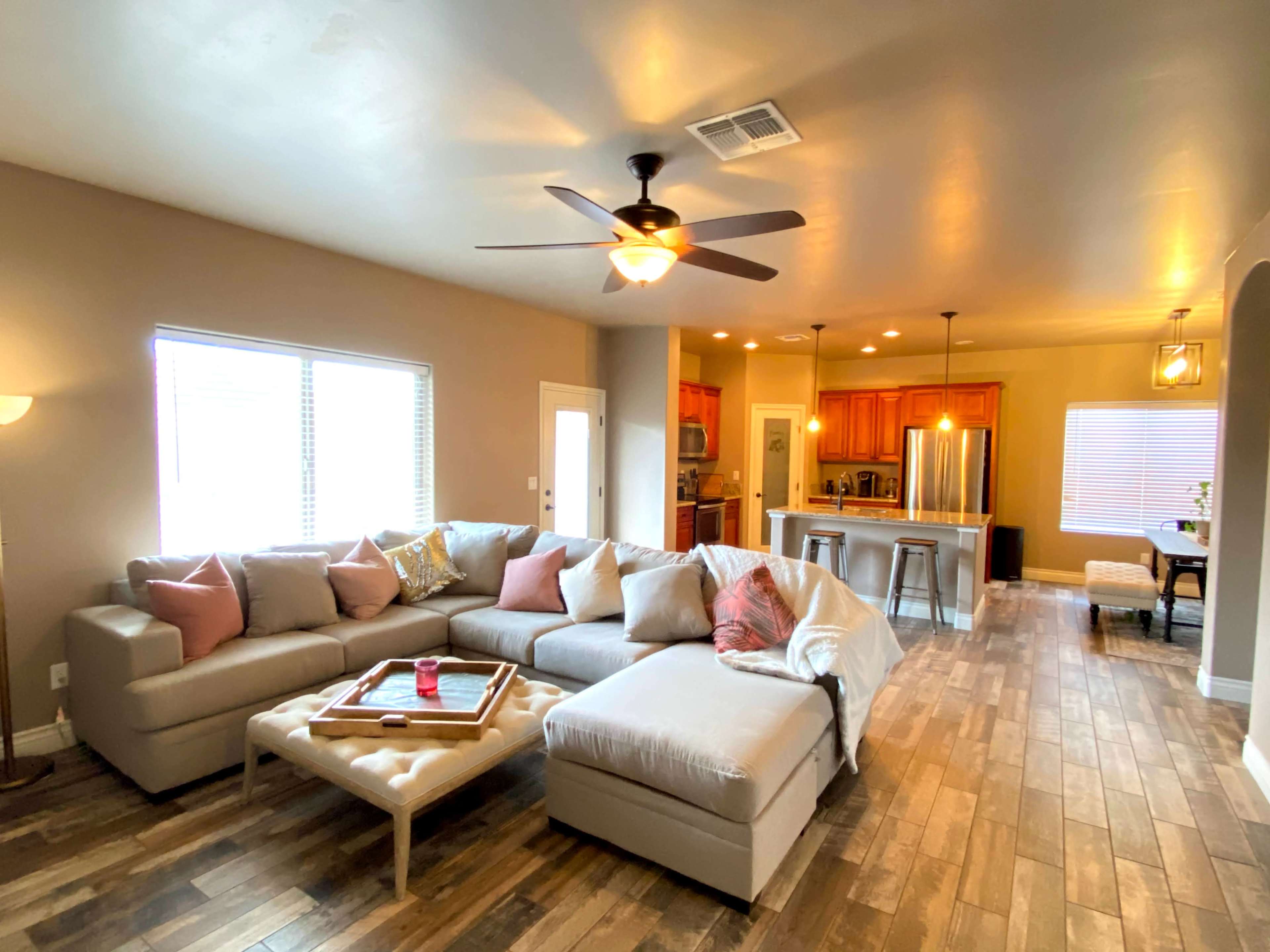 A spacious living room features a large sectional sofa, a coffee table, and an open kitchen area in the background with wooden cabinetry.