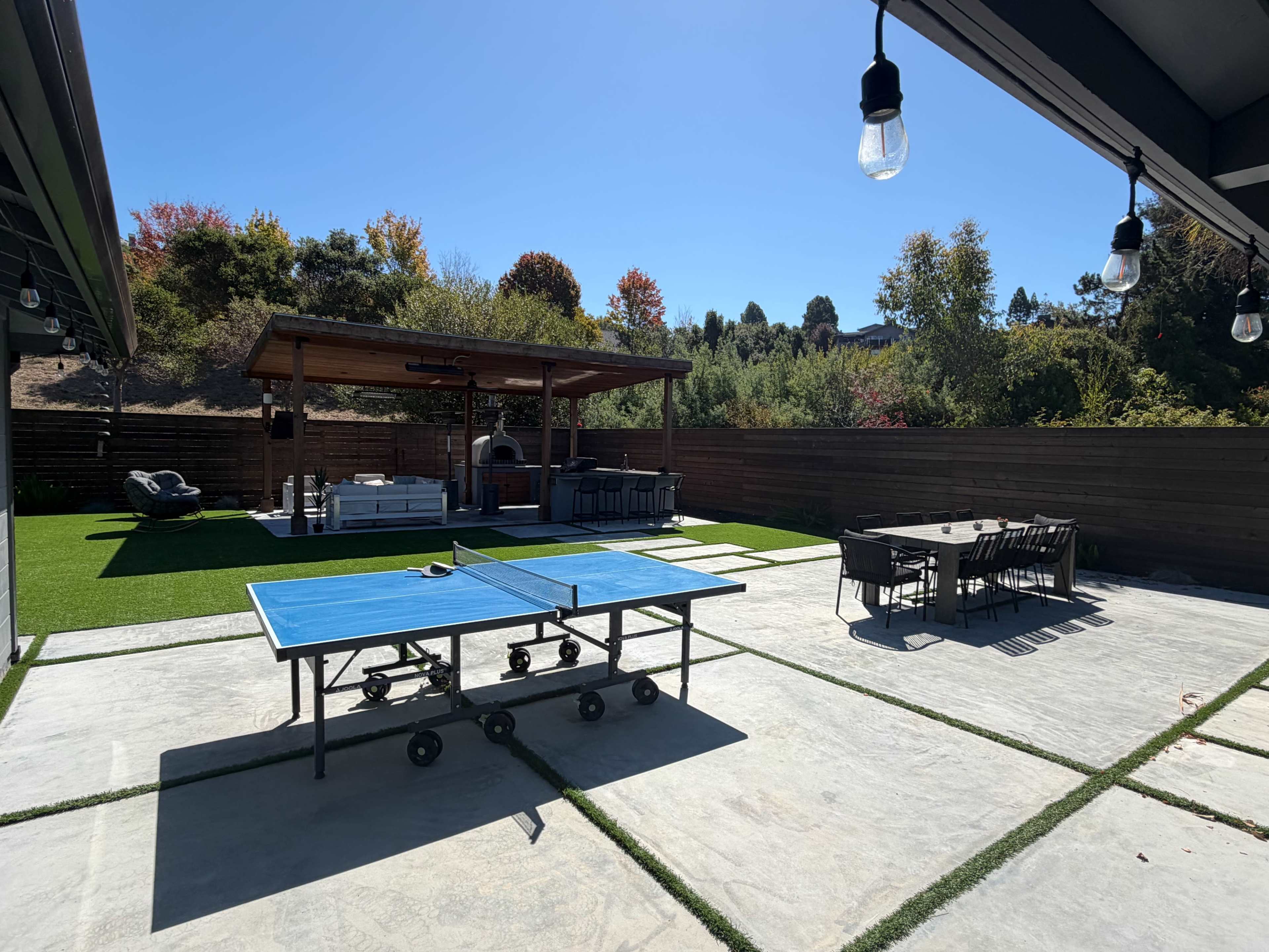 The image shows a backyard with a blue ping pong table, a covered patio area with outdoor seating, and a dining table, surrounded by greenery and a wooden fence.