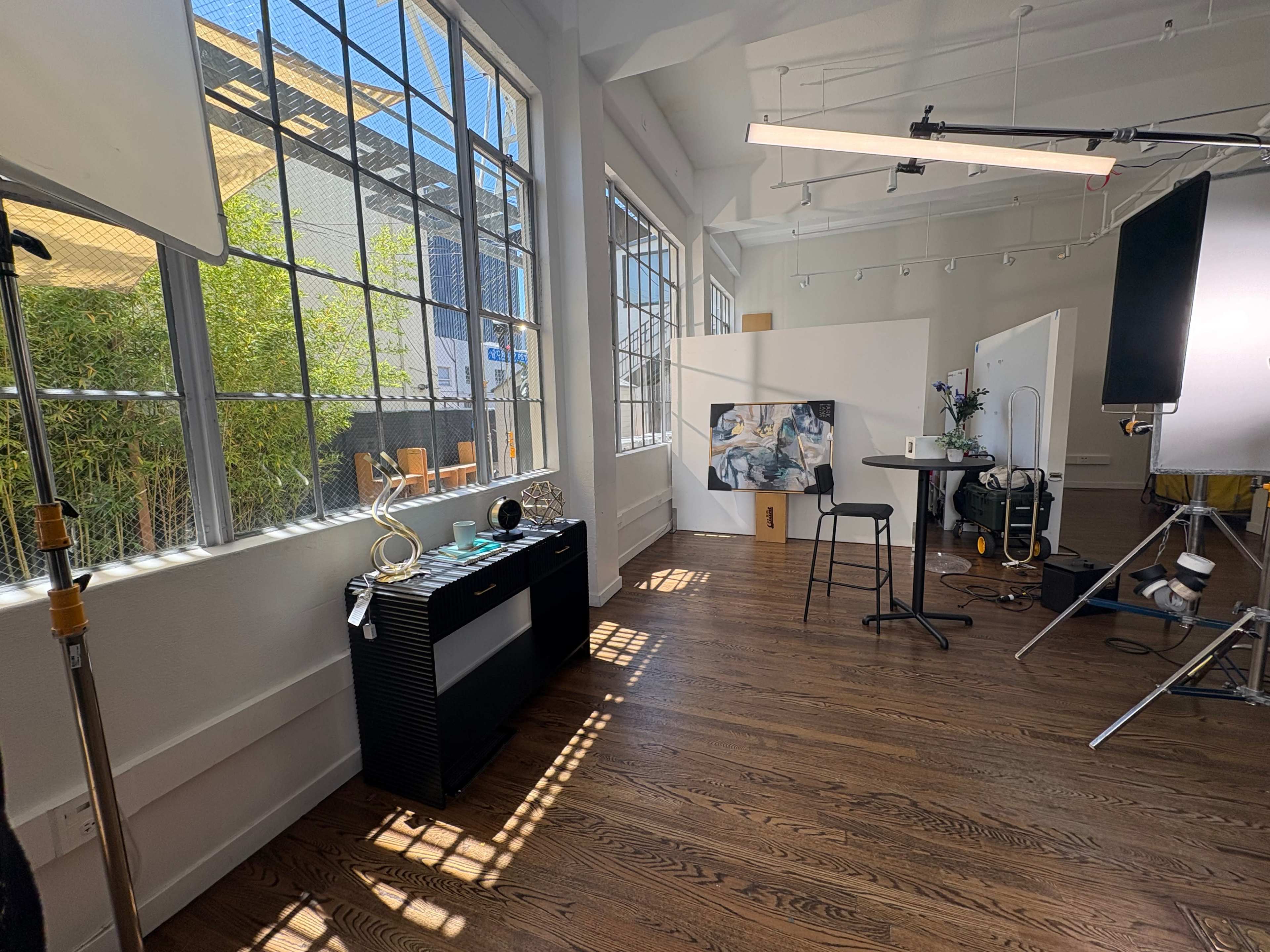 The image shows a bright studio space with large windows, a black console table, and a setup for photo or video production including a backdrop and lighting equipment.