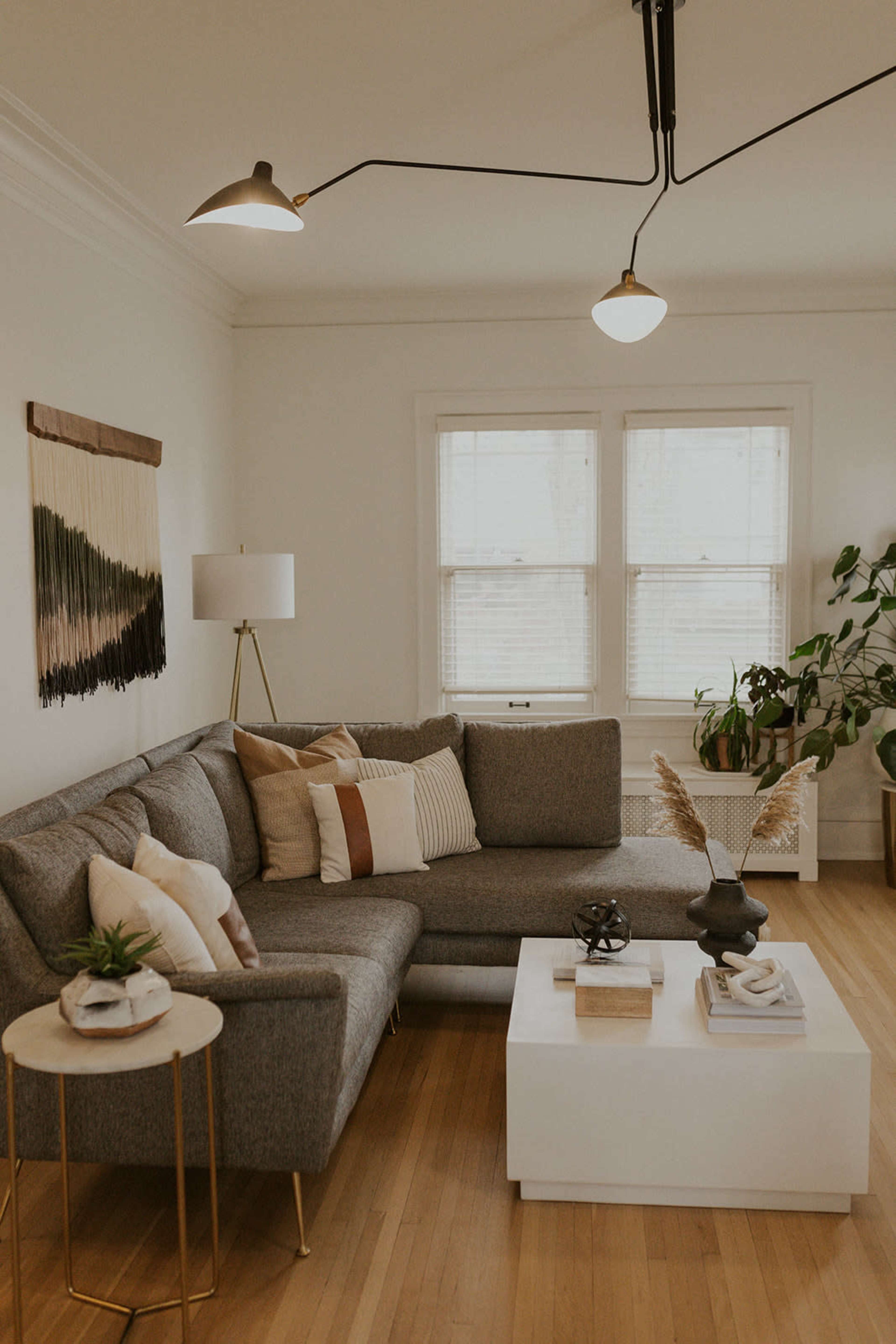 A modern living room featuring a gray sectional sofa with decorative pillows, a white coffee table with various decor items, and large windows allowing natural light in.