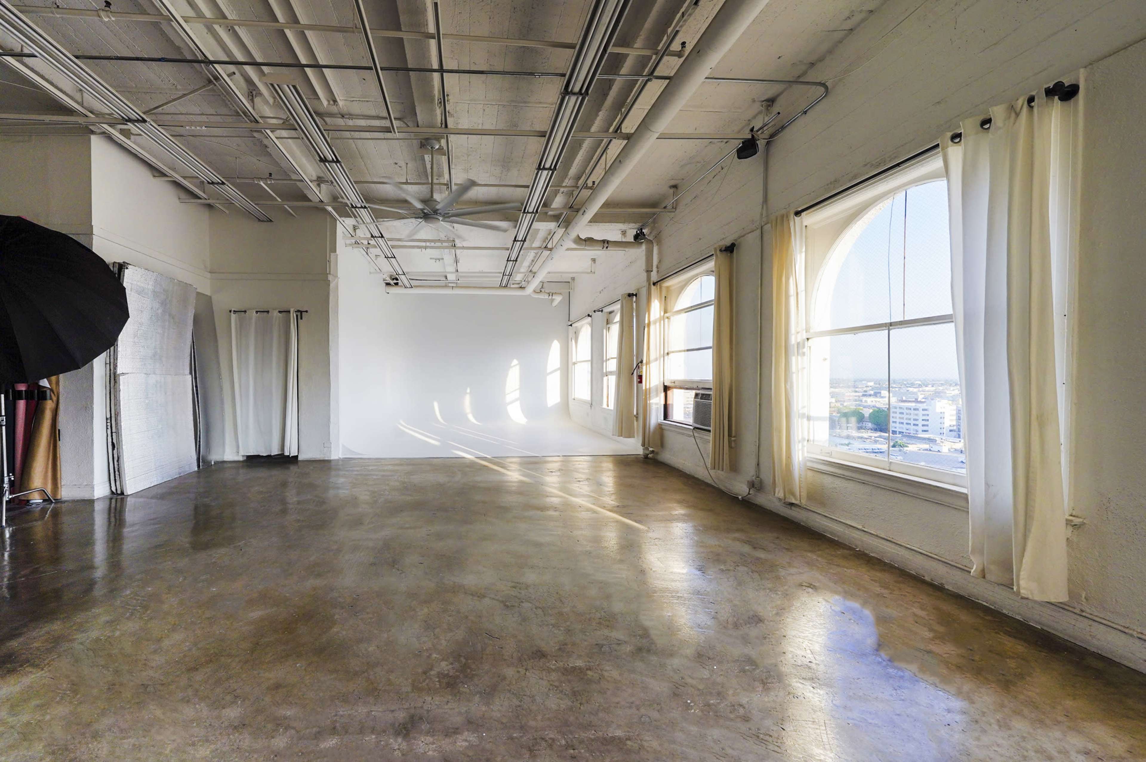 The image shows a spacious studio with large windows, concrete flooring, and a backdrop setup in the corner.