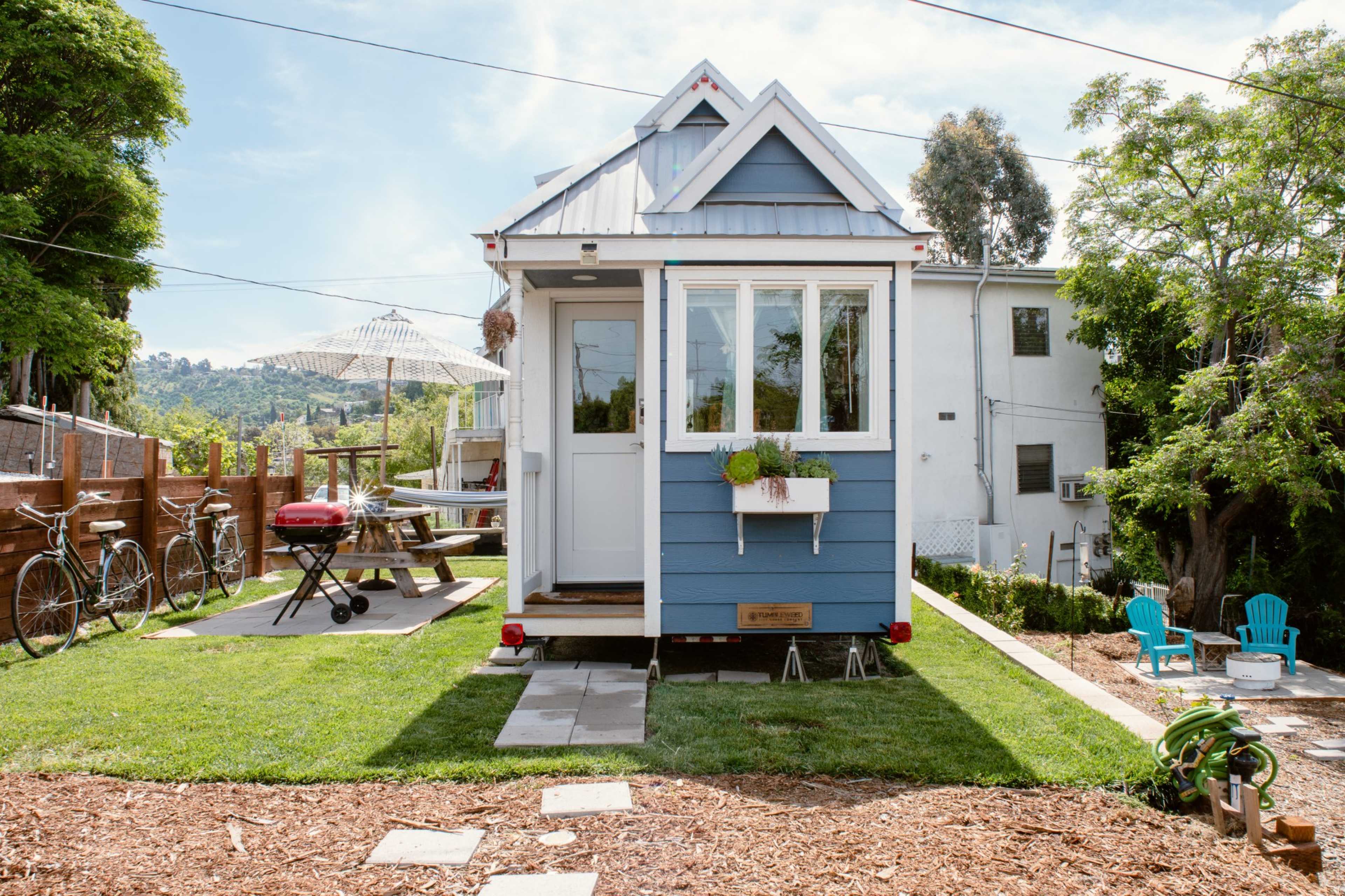 A small blue house with a peaked roof is set in a grassy yard, featuring a picnic table, barbecue grill, and bicycles nearby.