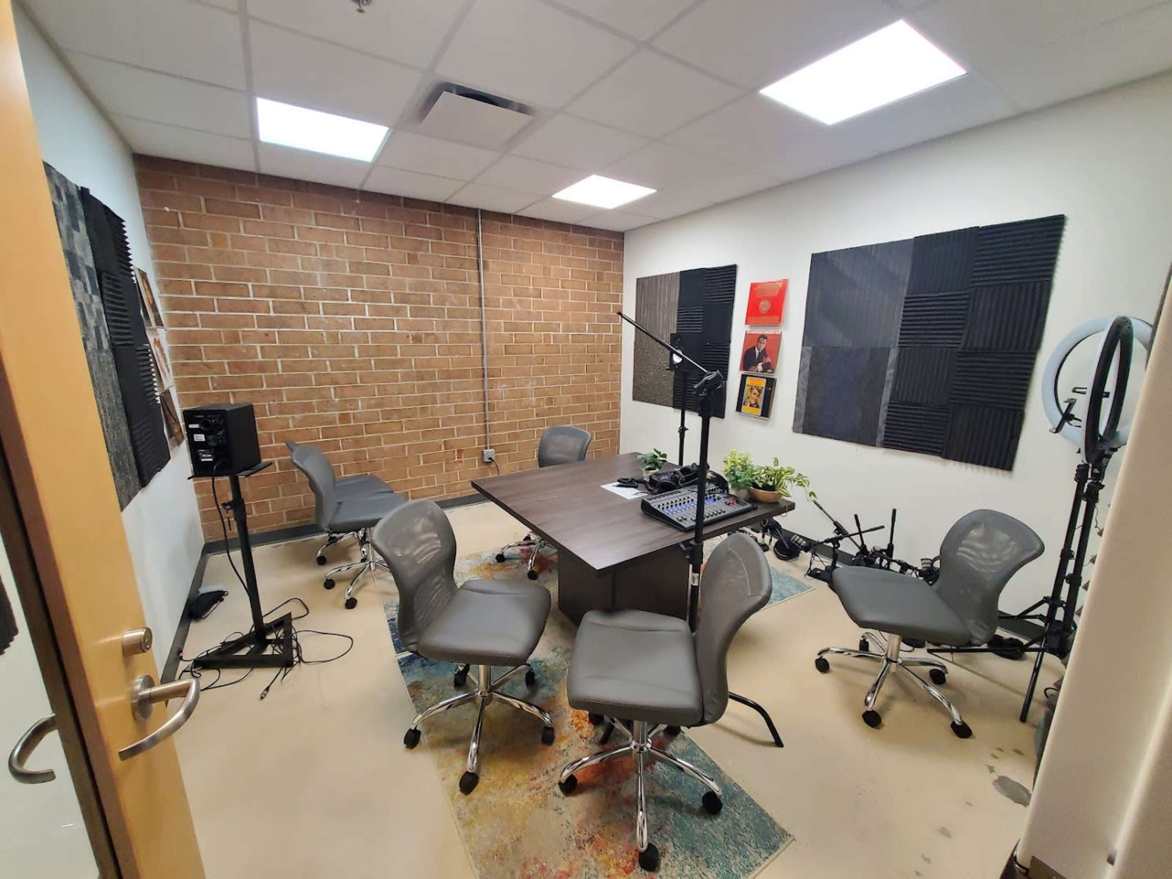 A small audio recording studio is furnished with a central table, several ergonomic chairs, soundproof wall panels, and recording equipment.