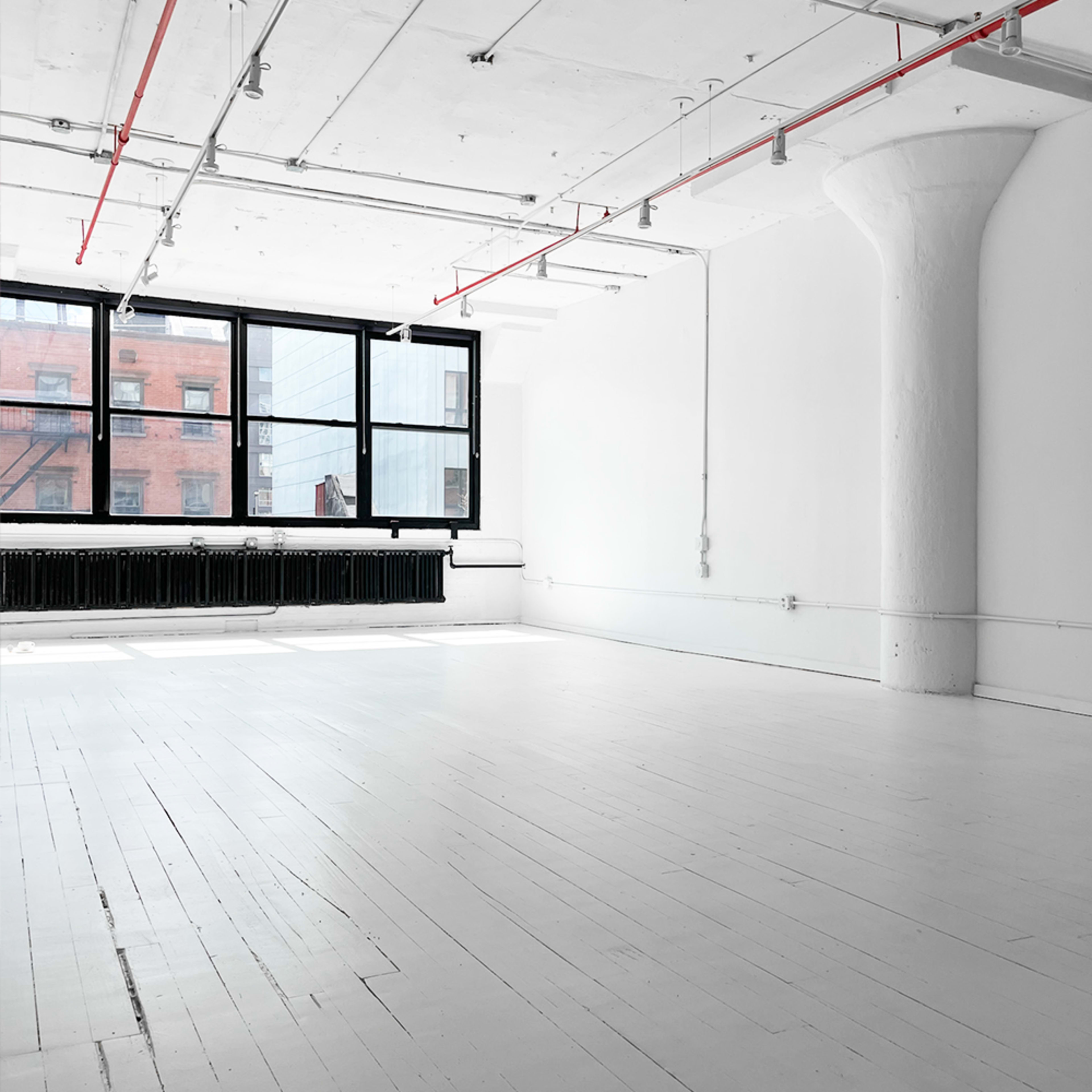 A spacious, minimalist room with white walls and floors, large windows, and exposed pipes.